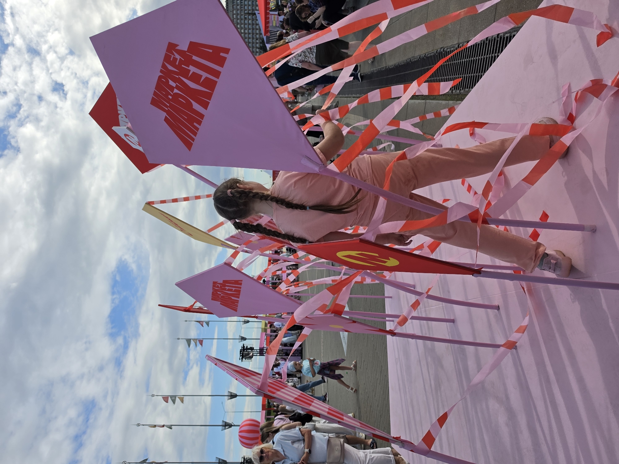 A young girl with braided hair, wearing a light pink t-shirt and pants, stands in the foreground of an outdoor public space. She is facing away from the camera, her back to the viewer, and her hands are positioned on a large, light pink diamond-shaped structure. This structure is adorned with red and white striped tape, and appears to be part of an art installation or event. Several other similar diamond-shaped structures, some red and some pink, are visible around her, also decorated with tape. In the background, a crowd of people are milling about, some walking, some standing. Further in the distance, a large red and white striped balloon is visible, along with flagpoles and tents, suggesting a market or festival. The sky above is partly cloudy. The scene appears to be taking place in an urban environment, possibly a public square or waterfront. Given the text visible on the pink diamond shape which reads "MARKET MARKETA" in red lettering, the location is likely the Nizhny Novgorod Fair (Nizhny Novgorod Yarmarka), a major exhibition and trade center in Nizhny Novgorod, Russia.