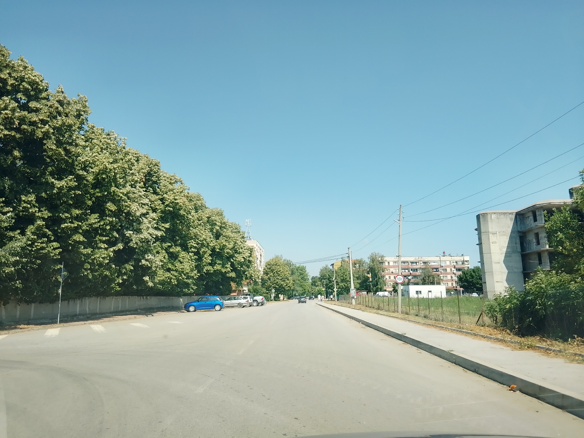 The image shows a wide, paved street in Ihtiman, Bulgaria, under a clear blue sky. On the left side of the street, a line of mature trees with green foliage and white blossoms forms a dense canopy along a concrete wall. Several cars are parked along the side of the road, including a bright blue hatchback. A pedestrian crossing is visible on the asphalt.
The street extends into the distance, with buildings and more trees lining both sides. On the right side, a sidewalk runs parallel to the road. Further down the street, there are apartment buildings with multiple stories. An unfinished concrete structure, possibly a building under construction, is also visible. Power lines crisscross the sky above. A circular road sign with "10T" in red is mounted on a pole. The overall environment appears to be a typical urban or suburban street scene on a sunny day.