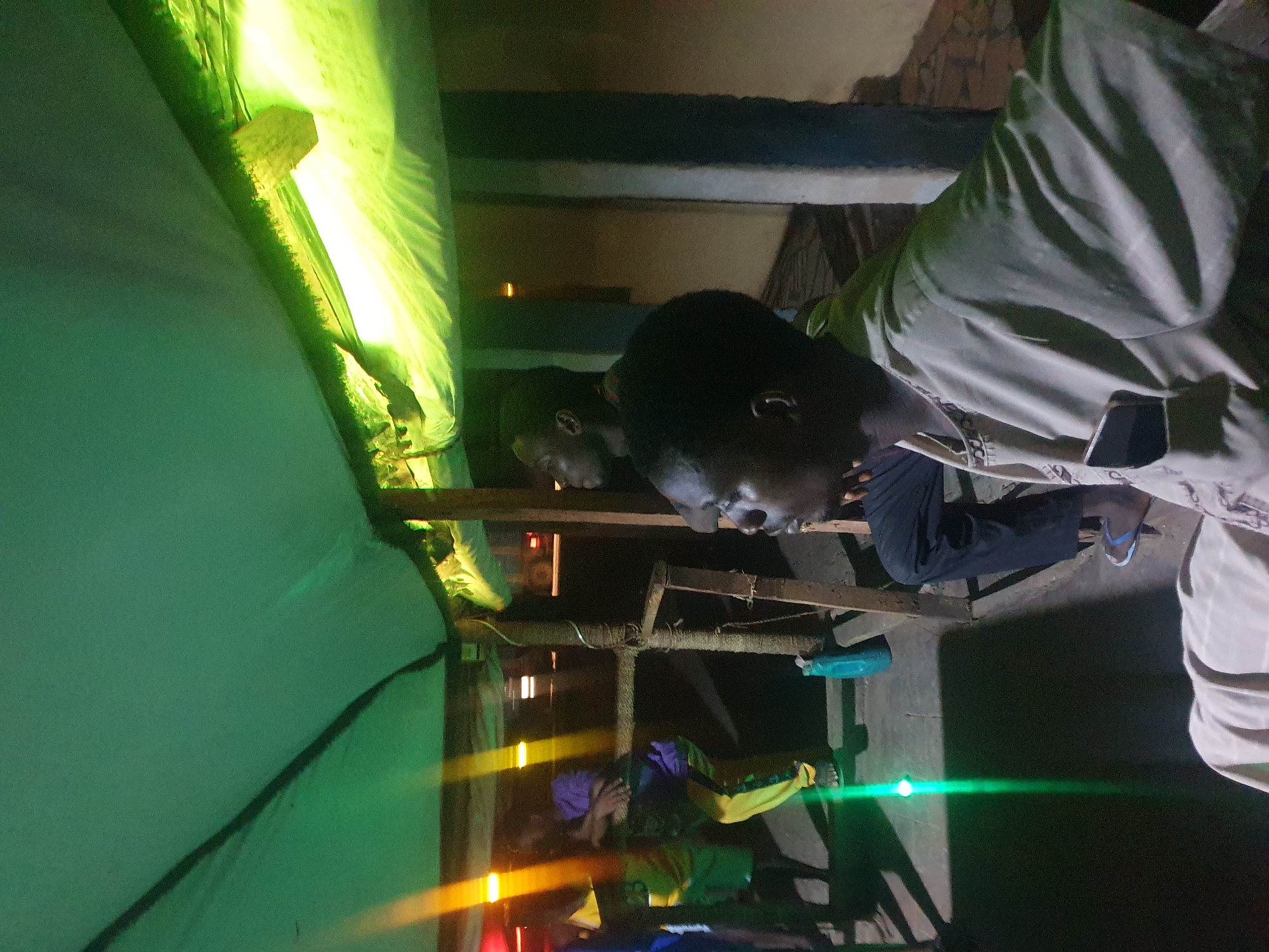 Two men are looking down, possibly at another person lying down on a bunk bed beneath them. The scene appears to be indoors, potentially inside a dormitory or a similar accommodation, possibly at night, evidenced by the dark lighting and the light stripes. One of the men is wearing a light-colored shirt. A mobile phone is visible on his clothing. A green tarpaulin is also visible. There is a bottle of water on the bed. The location is unknown without additional evidence, despite the prompt mentioning Lokoja, Nigeria.