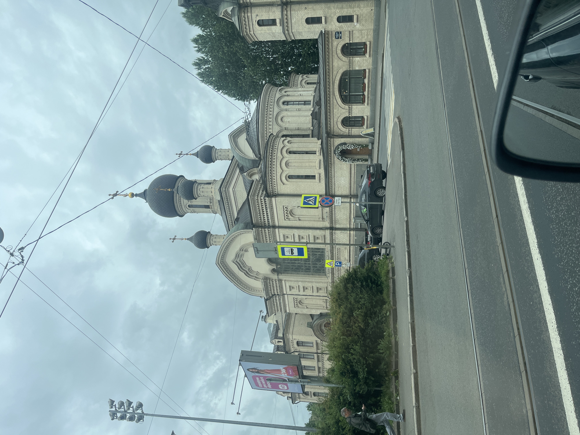 This media file depicts a view from a vehicle, looking towards a large, ornate church with multiple domes and crosses, typical of Russian Orthodox architecture. The church building is constructed of light-colored brick and features arched windows and decorative stonework. Overhead, a network of power lines crisscrosses the sky.
In the foreground, a road with tram tracks runs parallel to the church. Several cars are visible on the road and parked along the side. A pedestrian crossing is marked on the asphalt. A person is seen walking on the sidewalk, partially visible at the bottom of the frame. Adjacent to the church, a green hedge and a billboard advertising a product with an image of a woman in a pink dress are present. Traffic signs, including a bus stop sign and a pedestrian crossing sign, are also visible.
The sky is overcast and cloudy, suggesting a possibly cool or temperate day. The lighting indicates it is daytime, but the specific time of day is not clearly discernible. The overall scene conveys a sense of urban life in Saint Petersburg, Russia, with a blend of historical architecture and modern infrastructure. No specific emotions are conveyed, and the activity appears to be a normal street scene. The text on the billboard is not fully legible but includes Cyrillic script.