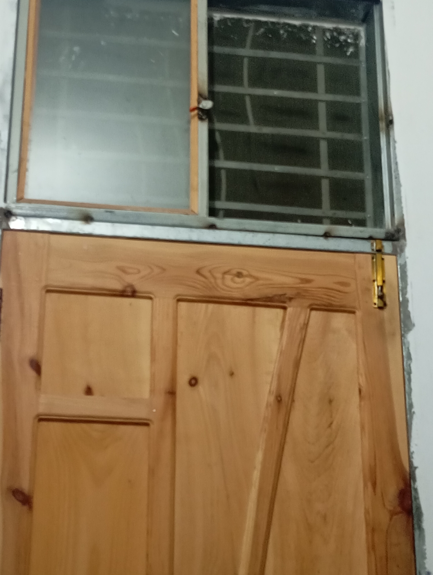 The image shows a wooden door with a small window above it. The window is covered with metal bars. There is a yellow latch on the right side of the door. The scene appears to be an interior shot, likely of a residential or commercial building. The image is taken from a low angle, looking up at the door and window. There are no people visible in the image. The environment is dimly lit and appears to be somewhat cluttered. The specific location in Lassan, Pakistan, cannot be determined from the image alone.