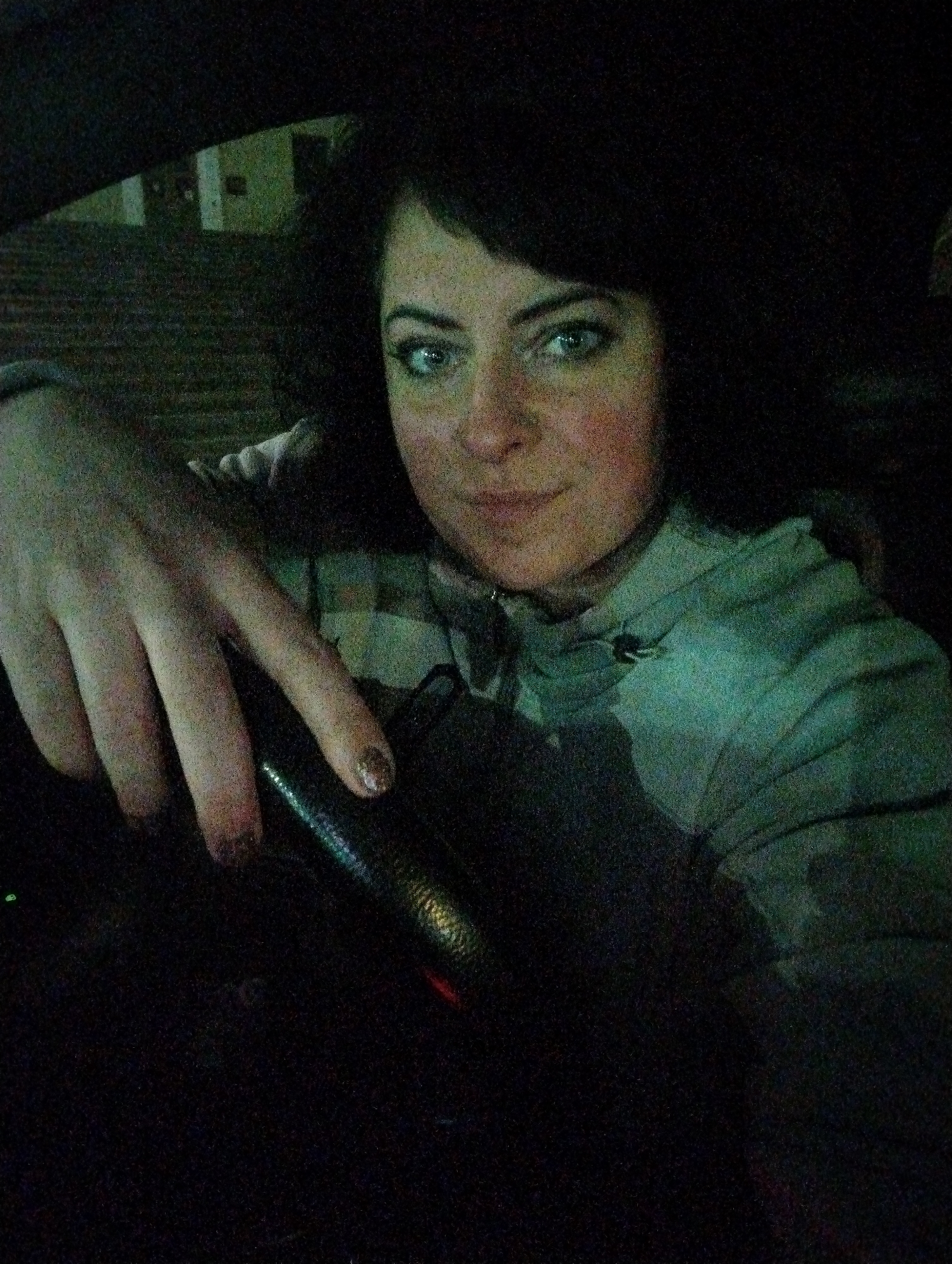 A woman is seated in the driver's seat of a car, presumably in Votkinsk, Russia, although no direct evidence of this is visible. The interior is dimly lit, with the woman's face and upper torso visible. Her left hand rests on the steering wheel. The background shows a blurry view of a building outside the car window. The image is dark and grainy, typical of low-light conditions.