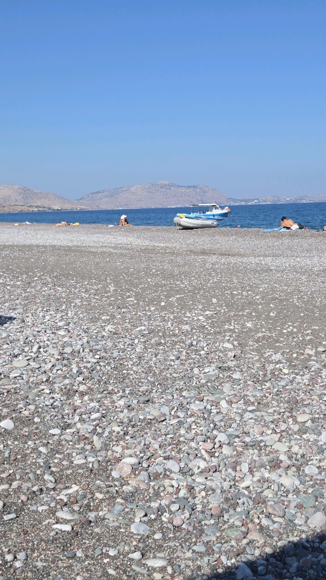 The image depicts a pebble beach in Gennadi, Greece, under a clear blue sky. The foreground is dominated by a textured expanse of grey, white, and reddish-brown pebbles. In the middle ground, several people are visible on the beach, some reclining and others sitting. The sea, a deep blue, stretches out to meet a distant, hazy coastline with hills. Two boats are in the water near the shore; one is a small, white and blue vessel, and the other is a gray inflatable boat. The scene conveys a tranquil atmosphere of a seaside vacation.