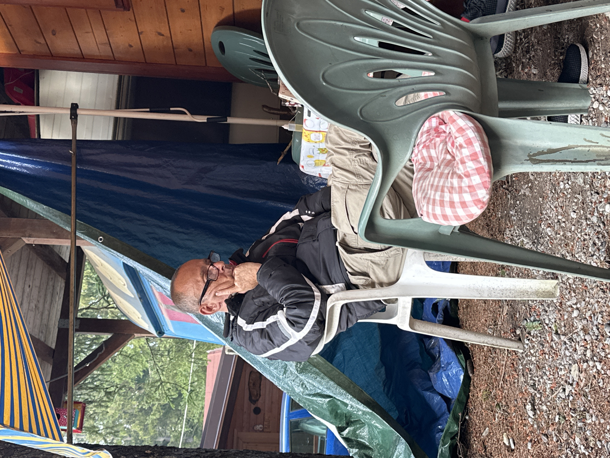An older Caucasian man wearing glasses, a black jacket with white stripes, and khaki pants is sitting on a white plastic chair. He has his right hand on his chin, looking down. He is seated outdoors, under a wooden structure with a blue tarp behind him. In front of him, on a small table covered with a checkered tablecloth, are a bottle of liquid and some papers. To his right, a green plastic chair with a red and white checkered cushion is visible. The ground is covered with gravel and wood chips. The background shows trees and a wooden building. The location is identified as Borno, Italy.