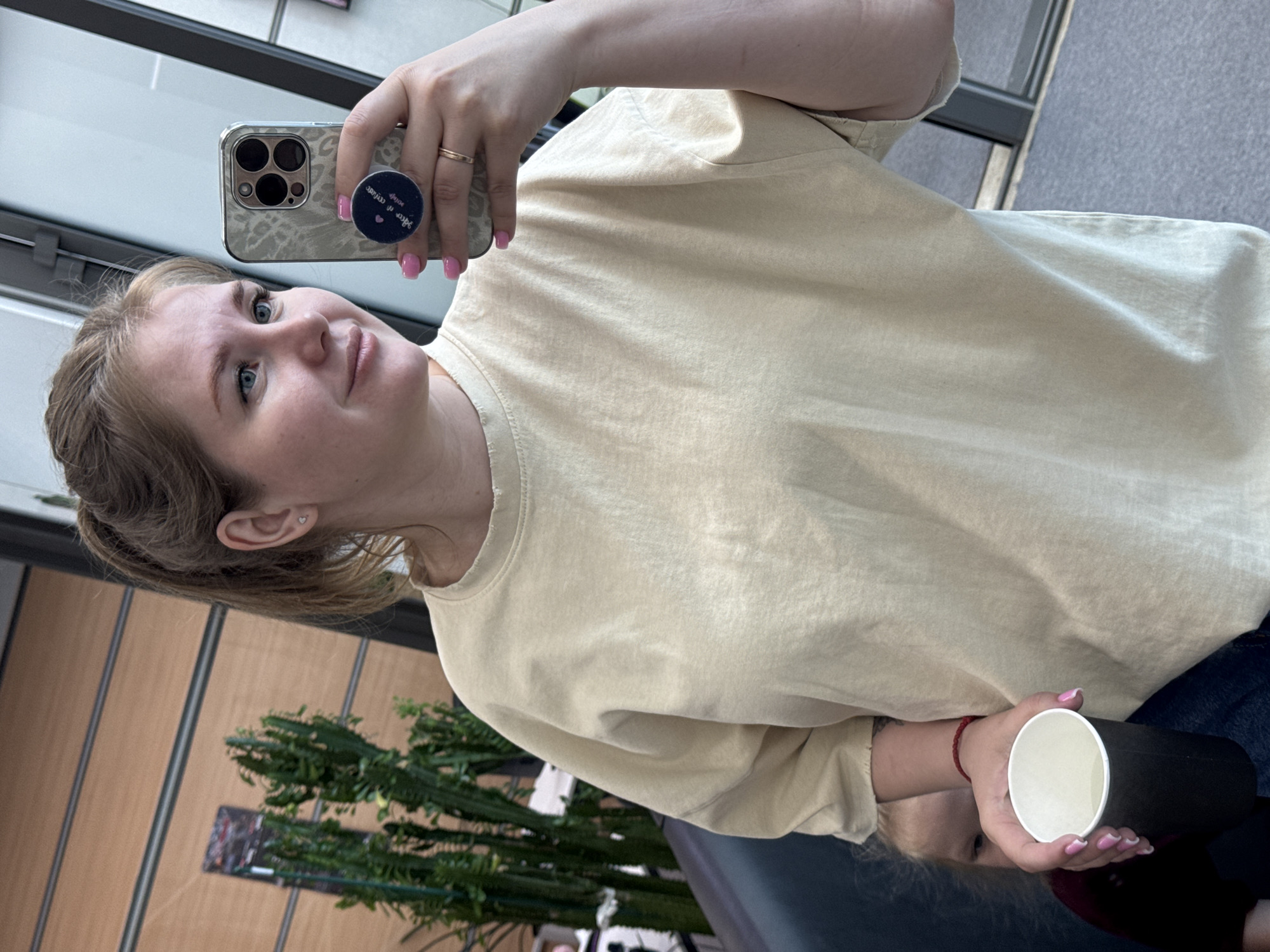 A woman is taking a selfie indoors. She is holding a smartphone with a patterned case and a pop socket. She is wearing a light beige t-shirt and a ring on her finger. She is also holding a paper cup with a black sleeve in her other hand. The background includes a wall with vertical wooden panels and a large green plant, potentially a cactus. The image appears to be taken in Perm, Russia, possibly in a café or office setting.