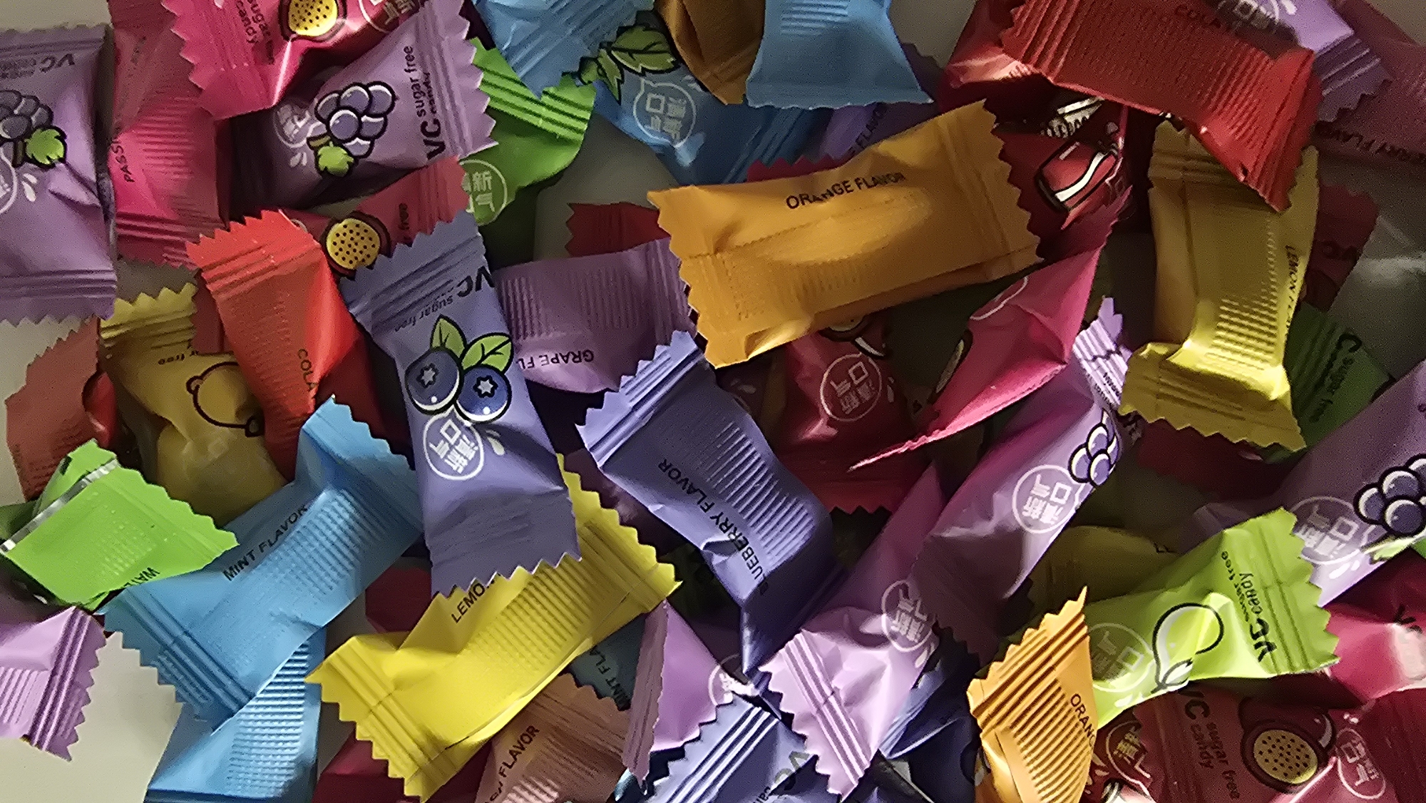 A close-up, top-down shot displays a chaotic pile of individually wrapped candies. The candies are a variety of colors including purple, pink, yellow, green, blue, and red. Many of the wrappers have text and graphics visible. Some of the text appears to be in English, with labels such as "MINT FLAVOR," "LEMON FLAVOR," "BLUEBERRY FLAVOR," "GRAPE FLAVOR," and "ORANGE FLAVOR." Other wrappers show images of fruits like blueberries and grapes, and some have Chinese characters. The wrappers are crinkled and have serrated edges. The overall impression is of a jumble of brightly colored sweets. The image does not contain any information or visual cues that would allow for the determination of a location within Chaikovsky, Russia.