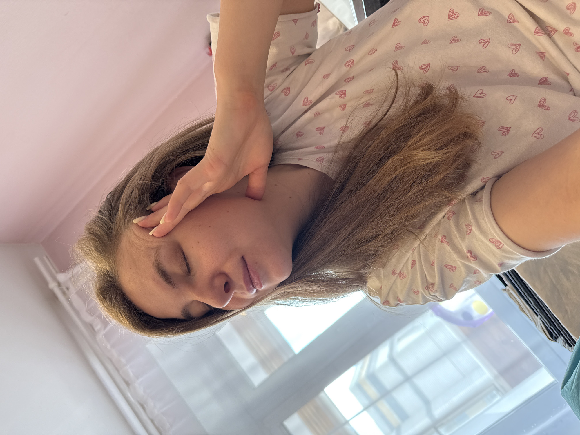 An indoor, overhead shot from Vladimir, Russia, shows a young woman with light brown hair, her eyes closed, seemingly resting or experiencing discomfort. The image is rotated 90 degrees clockwise. Her left hand is gently pressed against her temple, suggesting she might be tired, sleeping, or have a headache. She wears a light-colored t-shirt or pajama top patterned with numerous small pink hearts. Her long hair is visible around her head and shoulders.
The setting is a brightly lit room, indicating daytime, likely morning or afternoon, with clear weather outside. A large window, through which bright light streams, is visible in the lower right portion of the frame. The upper left shows a light pink wall or ceiling. No text is visible in the image. Her fingernails, painted a light shade, are discernible on her left hand. The overall mood is quiet and introspective, conveying either peaceful rest or a sense of weariness.