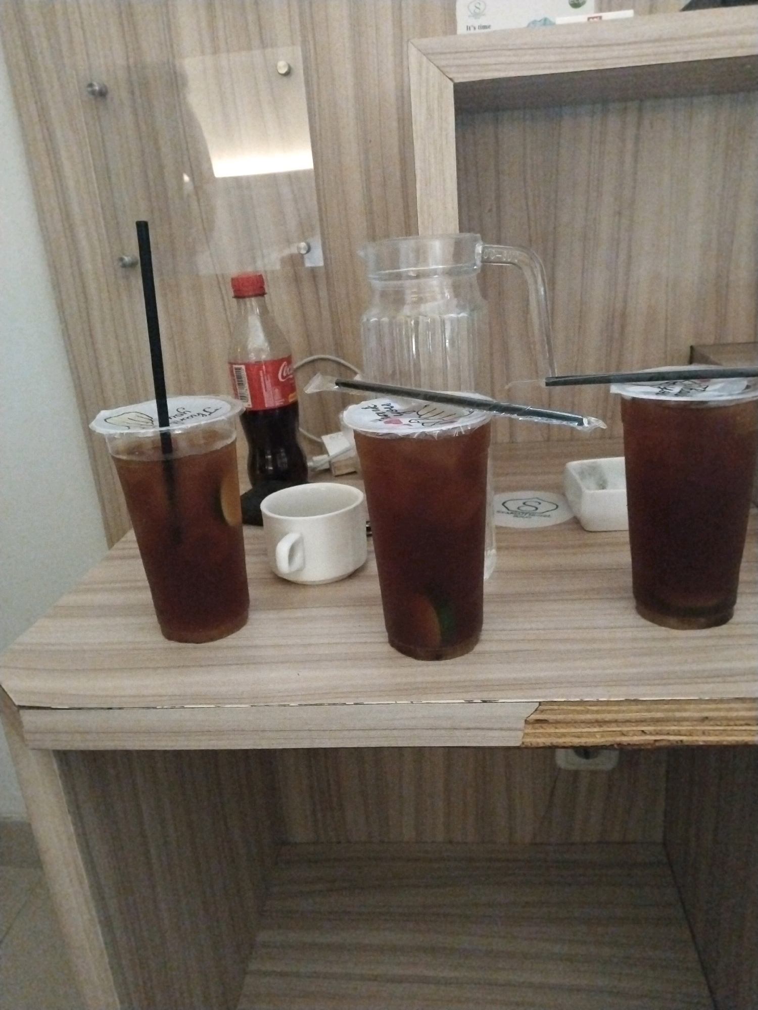 The image captures a close-up view of three iced tea drinks on a wooden table, positioned in what appears to be a hotel room or guesthouse. The drinks are in clear plastic cups with white lids and black straws. One drink is to the left, another in the middle, and a third to the right. A Coca-Cola bottle is visible behind the middle drink, and a small white cup sits next to it. A glass pitcher is partially visible in the background. The table surface is light-colored wood with a visible crack or damage on the front edge. The background features a wooden wall with some metal fixtures and a reflective panel with a light source behind it. The overall scene suggests a casual setting, possibly after a meal or during a relaxation period. There are no people visible in the image. The location is identified as Colomadu, Indonesia.