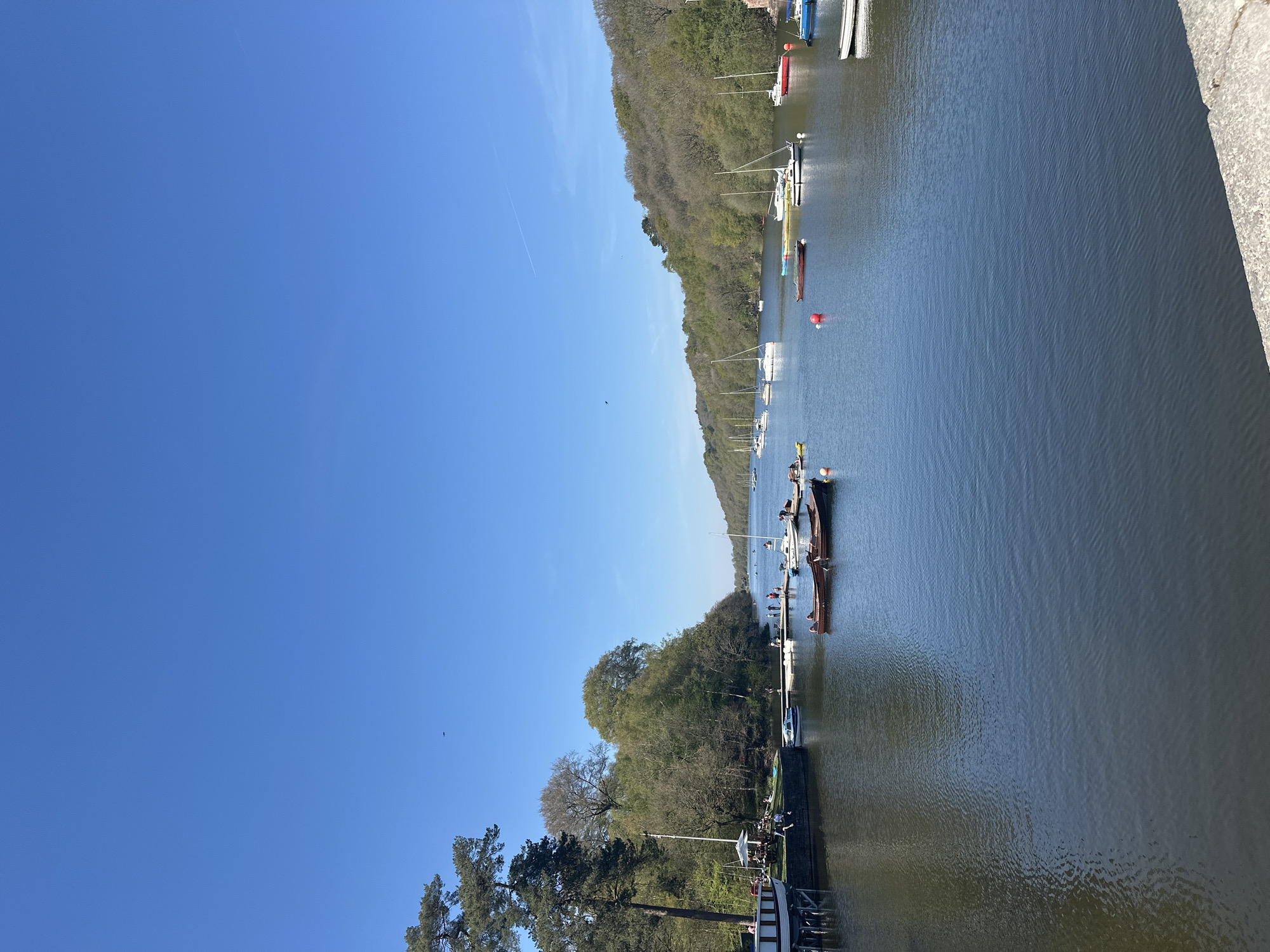 The image depicts a scenic view of a body of water, likely a river or lake, in Staffordshire Moorlands, United Kingdom. A line of boats and yachts of varying sizes is docked along the water's edge, indicating a marina or waterfront area. The opposite side of the waterway is lined with a dense forest, and the sky is a clear, bright blue. In the lower portion of the image, a path or walkway can be observed alongside the water, with people walking or sitting on benches. The overall atmosphere is calm and natural, suggesting a recreational area.