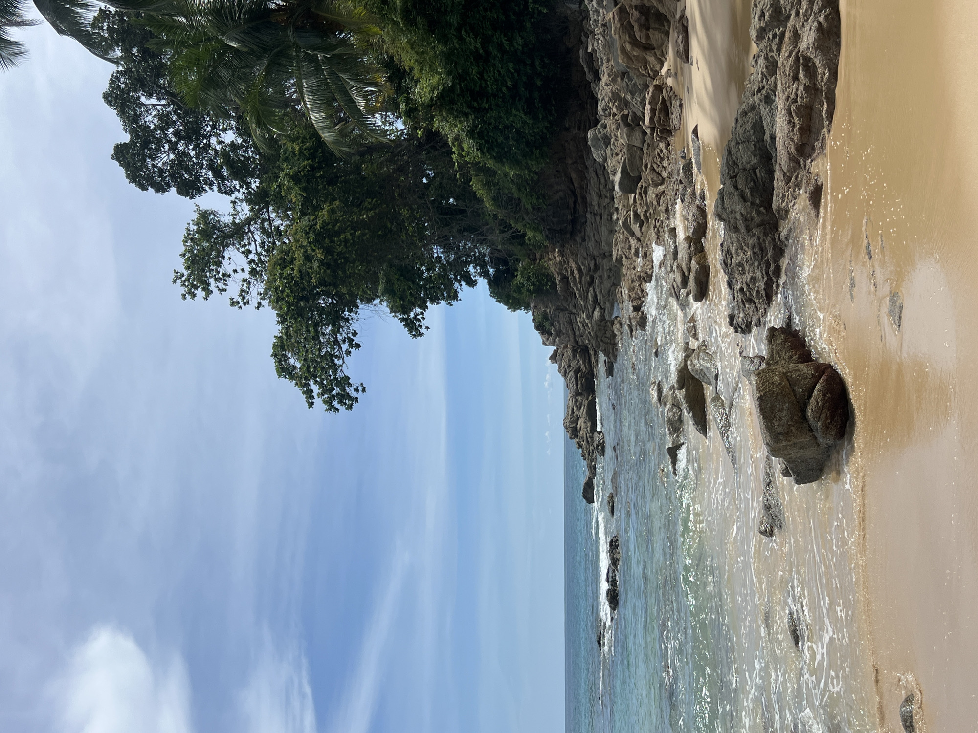 The image depicts a rocky beach in Choeng Thale, Thailand, on a clear day with a few wispy clouds. The foreground is dominated by a sandy beach, wet from the receding tide, with several large, dark gray rocks scattered across it. The ocean, a clear turquoise, laps gently at the shore, with small waves breaking over submerged rocks further out. To the right, a dense cluster of lush green trees and palm trees rises from a rocky outcrop, providing shade and a tropical feel. The overall impression is of a serene and natural coastal landscape.