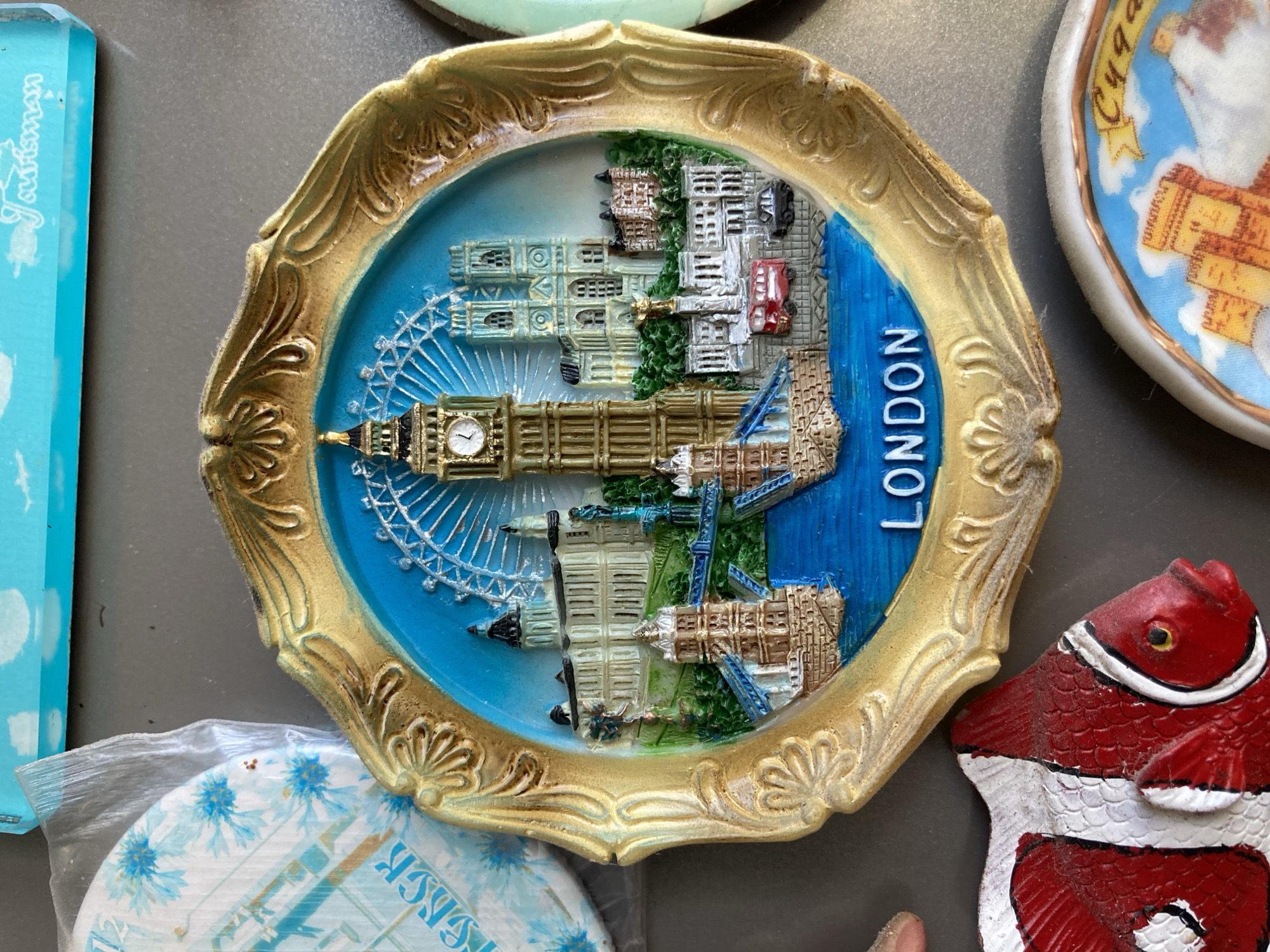 The image displays a close-up of a souvenir plate depicting London landmarks. The plate is framed with an ornate, gold-colored border. The central image showcases a miniature rendition of Big Ben, the London Eye, and Tower Bridge, set against a bright blue sky. A red double-decker bus and a black car are visible on the street below. The word "LONDON" is written in white letters at the bottom of the plate. To the left of the main souvenir, a partially visible blue and white patterned item, possibly another souvenir or packaging, is present. Below the London plate, a colorful ceramic fish souvenir, predominantly red and white with black accents, is visible. In the bottom right corner, another round souvenir with a castle illustration is partially visible. The background is a neutral, gray surface.