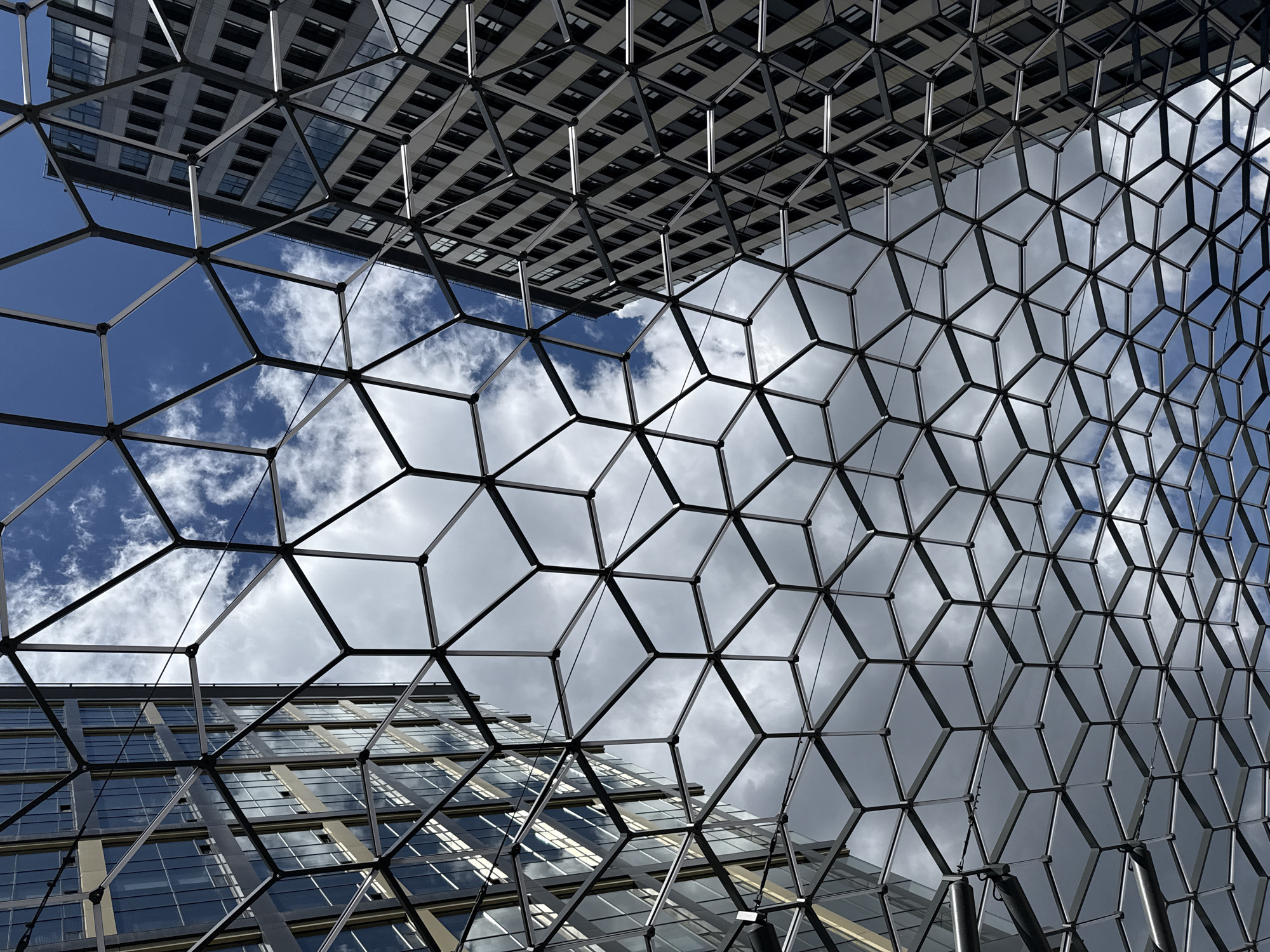 The image captures a low-angle perspective looking up at the geometric architecture of a modern building in Moscow, Russia. The foreground is dominated by a complex, hexagonal grid structure made of dark metal frames, which creates a tessellated effect across the frame. Through this grid, segments of a cloudy sky and the reflective facades of surrounding buildings are visible.
The sky is a mix of bright blue patches and overcast grey clouds, suggesting a dynamic weather pattern typical of a daytime scene. Sunlight reflects off the windows of the buildings, creating a sense of depth and highlighting the building's modern design. The buildings themselves appear to be office or residential complexes, characterized by their numerous windows and clean, linear construction.
The scene is devoid of people or any discernible activity, focusing entirely on the interplay of architectural forms, light, and sky. The geometric patterns of the foreground grid and the windows of the buildings create a sense of order and repetition. The overall impression is one of urban modernity and abstract design. There is no visible text in the image.