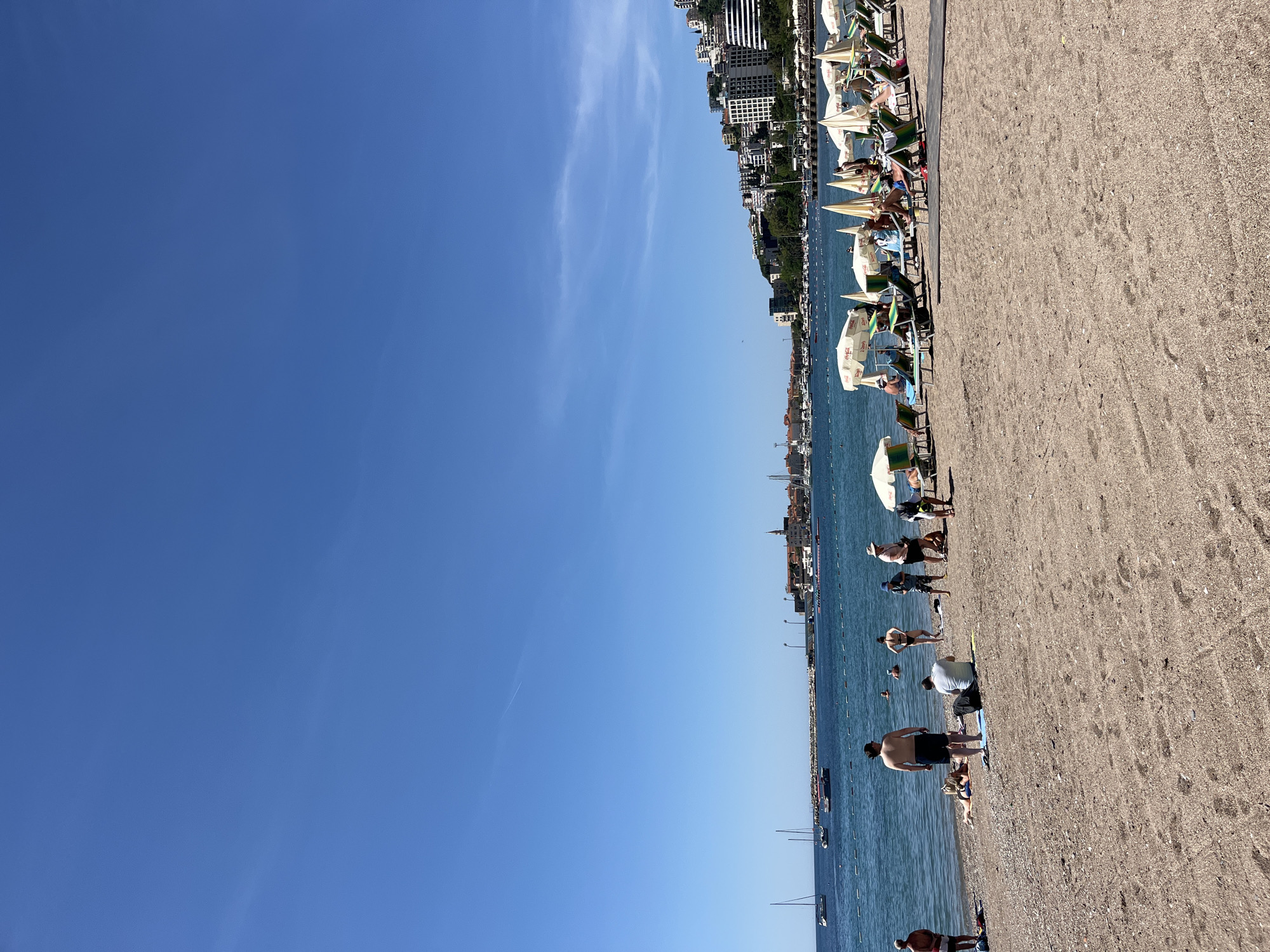 The image captures a beach scene in Budva, Montenegro, under a clear blue sky with few clouds. The beach consists of a sandy area and the Adriatic Sea. Along the shoreline, numerous beachgoers are seen engaging in activities such as sunbathing and walking. Beach umbrellas and sunbeds are arranged parallel to the shoreline. In the background, buildings and architecture characteristic of Budva are visible. A few boats dot the water.