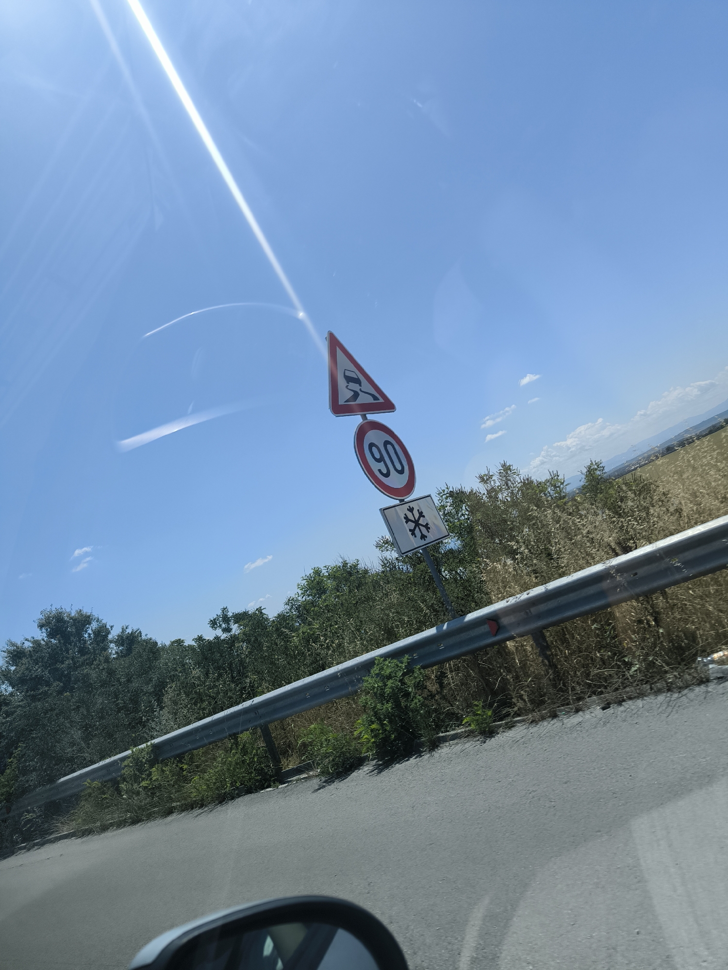 A road sign is mounted on a pole against a bright blue sky with a strong sun glare reflecting off a windshield. The sign displays three symbols: a triangular warning sign with a slippery road icon, a circular sign with the number "90", and a square sign with a snowflake symbol. Beyond the sign, a metal guardrail runs along the side of a paved road, bordered by dense green and dry brush vegetation. In the distance, rolling hills and fields are visible under the clear sky. The image appears to be taken from a moving vehicle, as evidenced by the reflection in what seems to be a car's side mirror in the lower foreground. The location is identified as Mirovo, Bulgaria.
