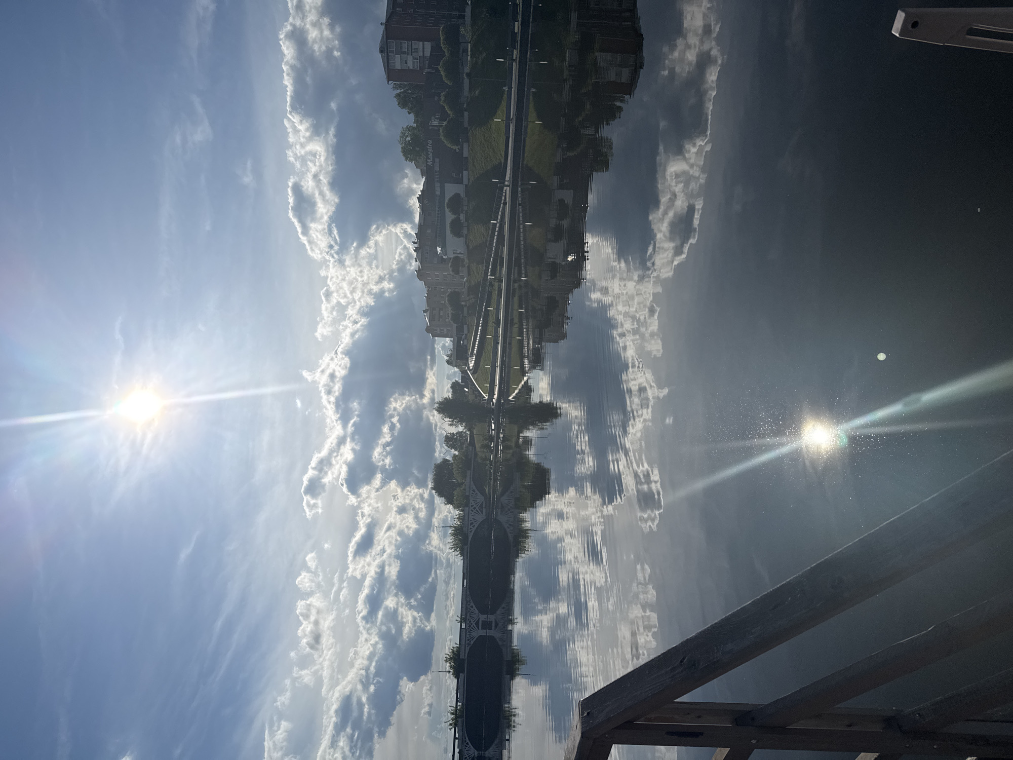 The image shows a reflection in water of a bridge in Votkinsk, Russia. The sky above is blue with cumulus clouds. A building with a sign is partially visible in the reflection. Part of a wooden structure is in the foreground. Two sun flares are also visible in the image.
