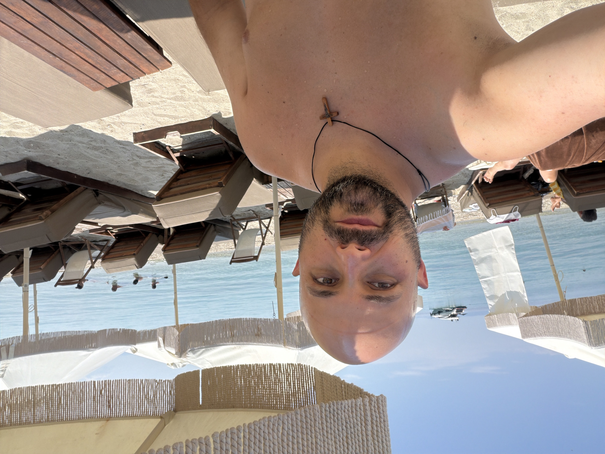 The image is a selfie taken at a beach in the city of Pallini Municipal Unit, Greece. The foreground features a man with a shaved head and a beard, wearing a black cord necklace with a wooden cross pendant. He is shirtless and appears to be wet, possibly from swimming. Behind him, a sandy beach is lined with rows of unoccupied lounge chairs and umbrellas. The sea is visible in the background with a few people swimming and a boat anchored offshore. The sky is clear and blue, indicating a sunny day. The overall scene depicts a relaxing day at a beach resort.