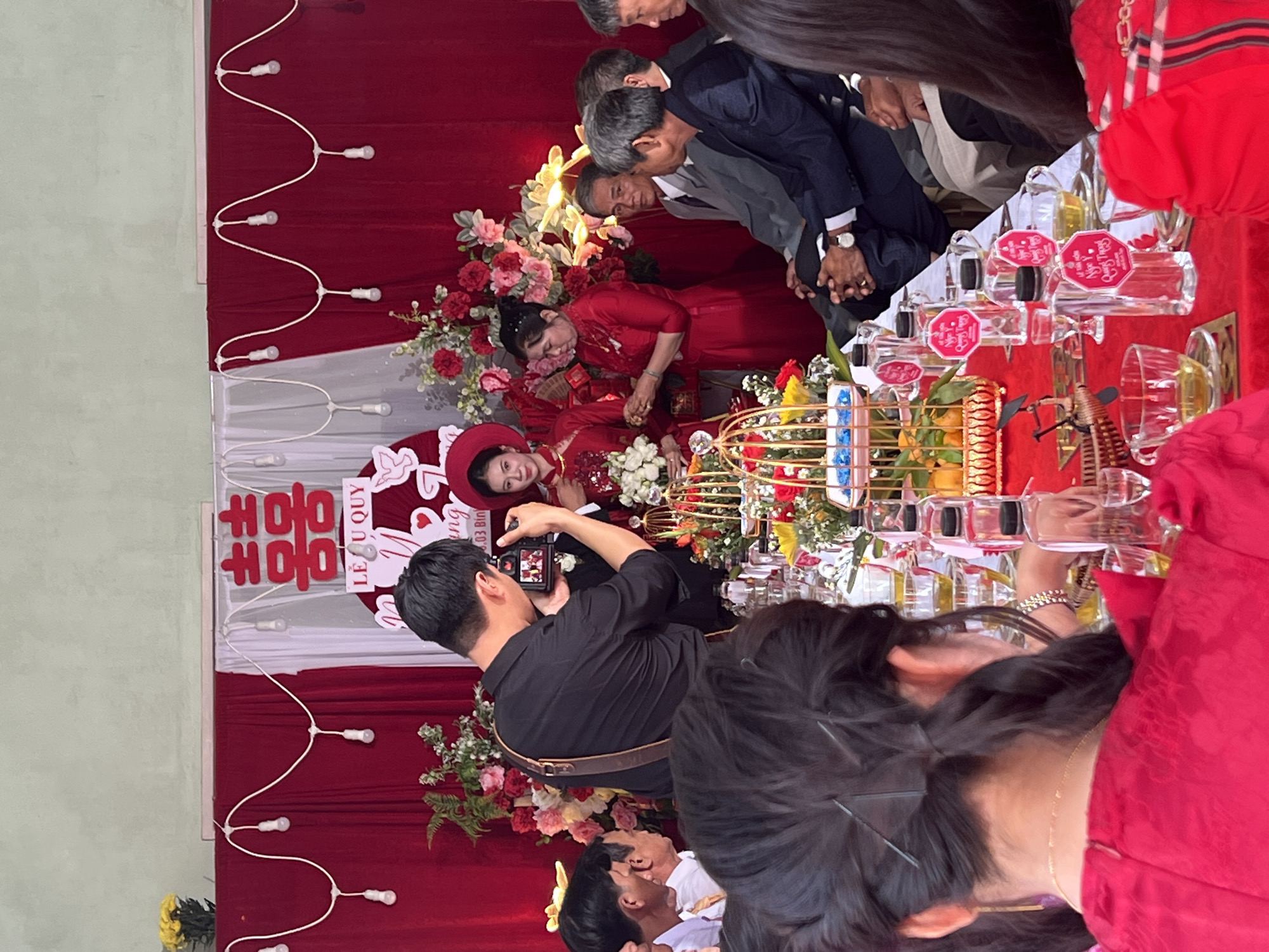 Captured in Tay Son Commune, Vietnam, the image depicts a wedding scene. A couple, adorned in traditional red attire, is the focal point, positioned in front of a backdrop featuring red fabric, lights, and floral arrangements. Several individuals surround them, some dressed in formal attire. One person, wearing a black shirt, is taking photos of the couple. A table is set with bottles, glassware, and an ornamental cage. The overall atmosphere suggests a celebration.