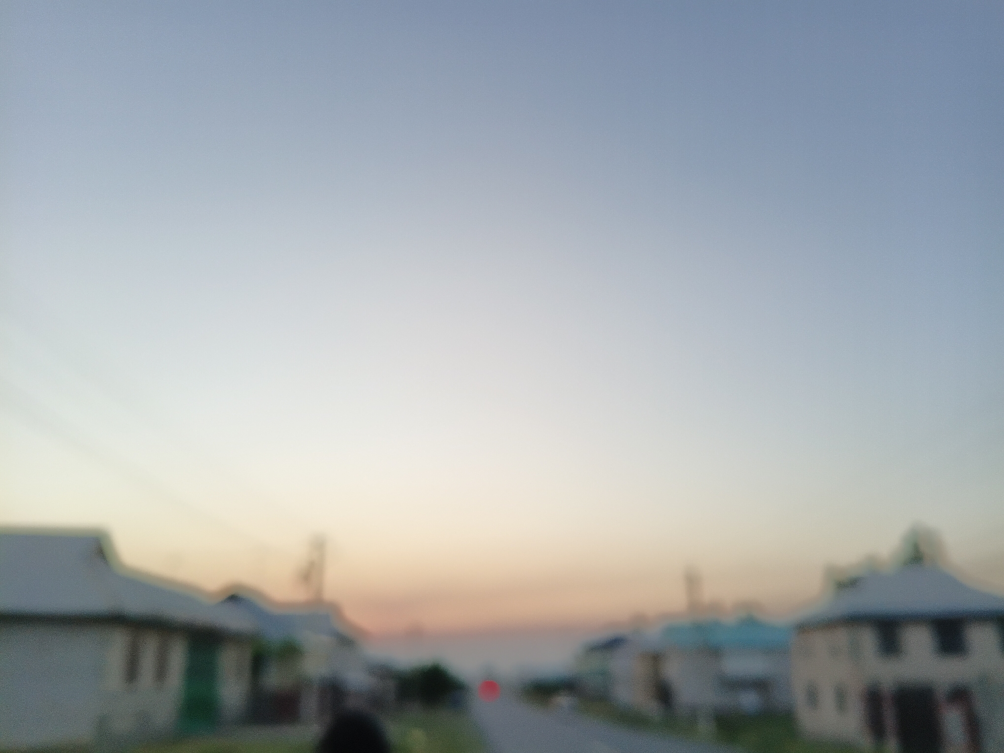 This is a blurry photograph of a residential street at dusk. The sky is a gradient of pale blue and soft orange, indicating the sun has recently set. The scene depicts a row of houses on either side of a road. The houses appear to be modest, single-story structures with peaked roofs, some painted in light colors, others with darker trim. There is a discernible road running through the center of the image, and a single red light, possibly from a vehicle, is visible in the distance. Two figures are partially visible in the foreground on the left and right sides of the image, their features indistinct due to the blur. Utility poles or thin trees are visible along the street. The overall impression is that of a quiet evening in a community.