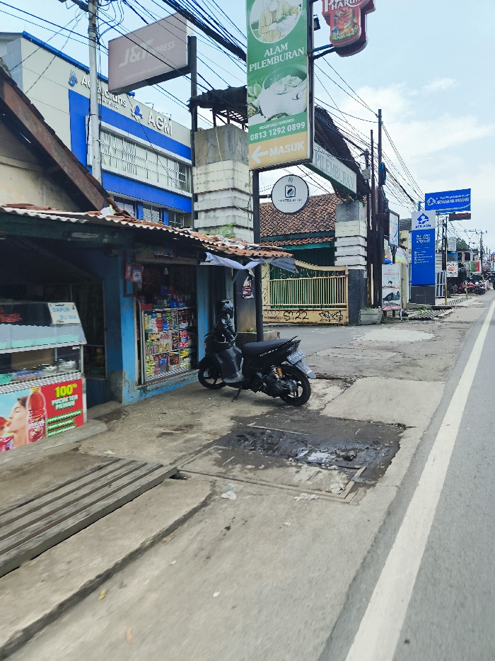 The image shows a street in Ngamprah, Indonesia, with a small shop on the left side of the road and a motorcycle parked in front of it. There is a sign above the shop that says "J&T Express". The shop's front is painted in blue and white. Behind the shop is a building with a blue and white sign. On the right side of the street is a sign that says "Alam Pilemburan" with a phone number and an arrow that points to the left. There is a white line down the middle of the road. The road is paved and the sidewalk is covered in concrete slabs. A small, green, leafy tree can be seen in the distance. The sky is a light blue with white clouds.