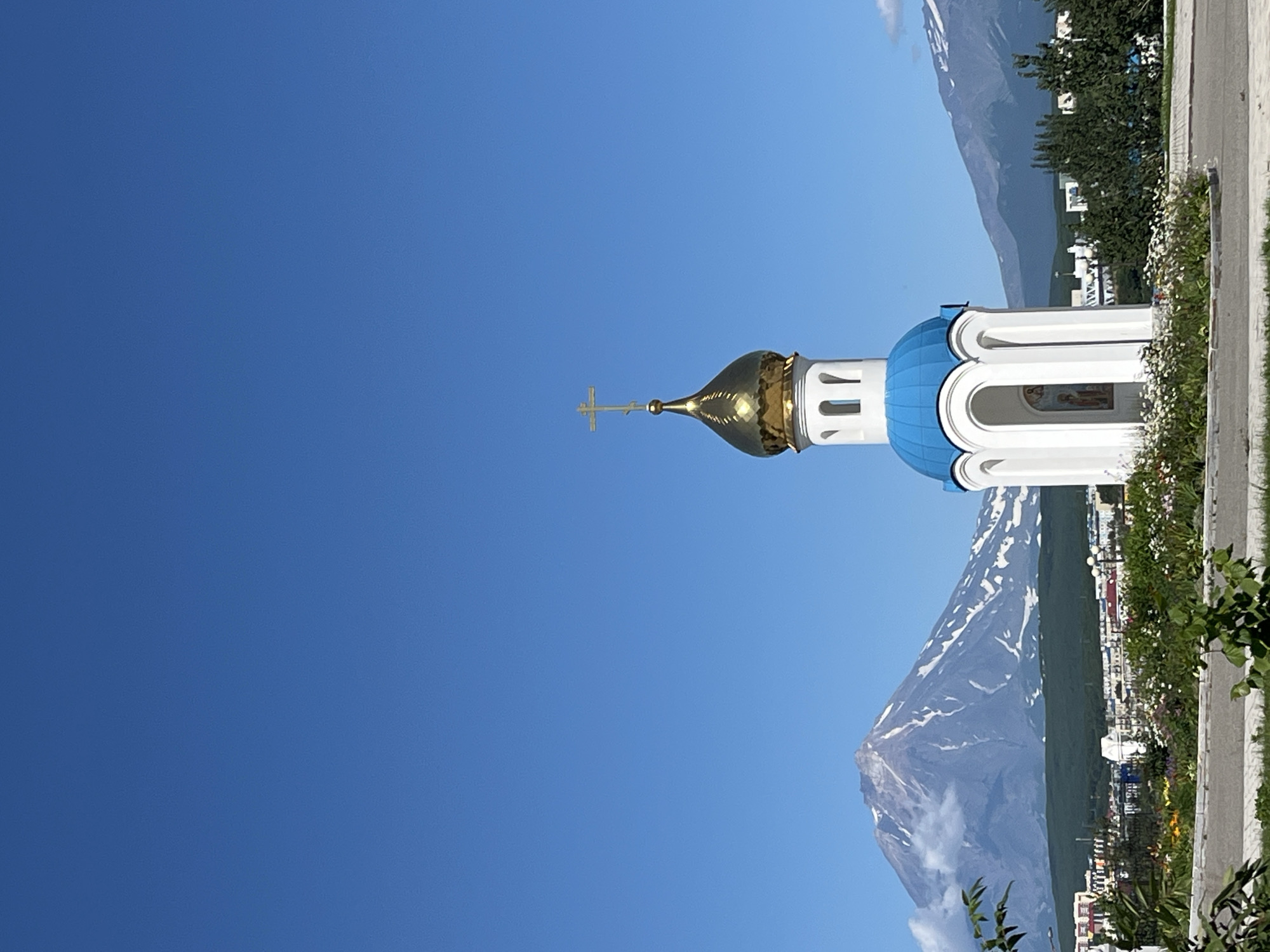 A clear, bright, sunny day with a deep blue sky over a landscape featuring a small white chapel with a golden dome and a blue roof. In the background, a large, snow-capped volcano dominates the horizon, with clouds partially obscuring its peak. A town is visible nestled at the base of the volcano, with a mix of buildings and greenery. The foreground shows a paved area and some flowering bushes. The scene is likely Petropavlovsk-Kamchatsky, Russia, given the prominent volcano, known as Avachinsky Volcano, which is a landmark of the region.