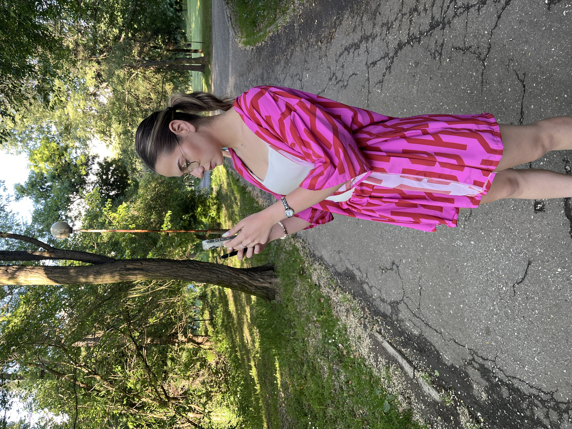 A woman stands on a cracked asphalt pathway in a park, looking at a cell phone in her hands. She is wearing a pink and red patterned dress over a white top, glasses, a watch, and a bracelet. The park features trees and grassy areas. It is not possible to tell if this image was taken in Blagoevgrad, Bulgaria.