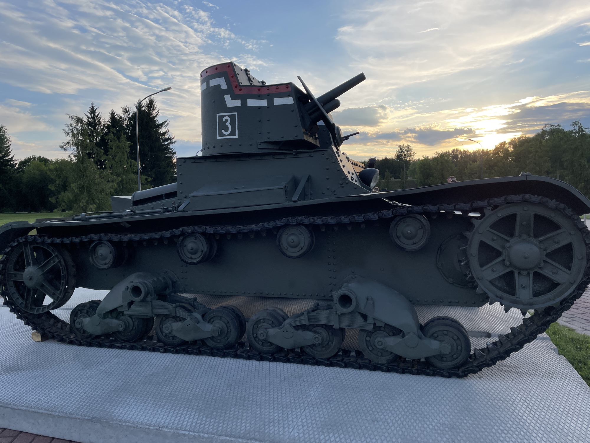 The image depicts a Soviet-era tank, likely a T-26 light tank based on its design, displayed outdoors on a paved platform. The tank is painted dark green and exhibits signs of wear and tear, suggesting it is a historical exhibit. The turret features a prominent gun and a marking of the number "3" within a square, along with red and white stripes. In the background, there are trees, a street lamp, and a cloudy sky with the sun setting on the horizon. The tank is positioned at the Museum of Fortification in Brest, Belarus, as part of its outdoor exhibit. No people are clearly visible interacting with the tank.