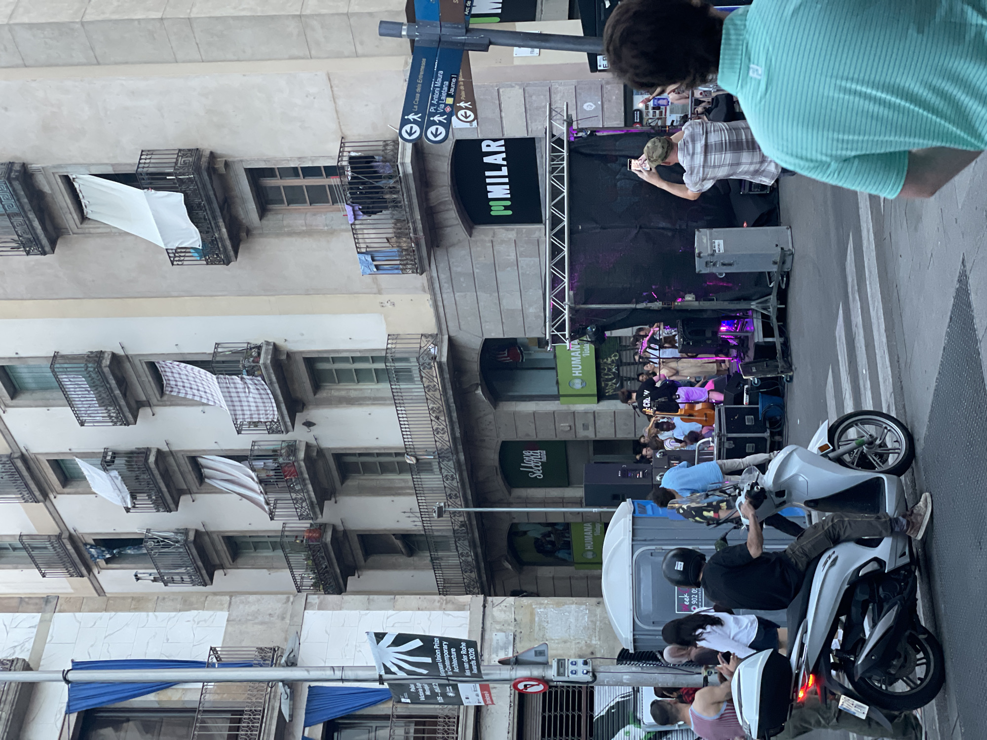 The image depicts a street scene in Barcelona, Spain, likely in a commercial or public area with a large building facade. The building features multiple balconies with wrought iron railings and various items hanging from them, including laundry and awnings. On the ground level, a stage has been set up with musical equipment, and a band appears to be performing. A crowd is gathered in front of the stage, with some people watching the performance and others engaged in activities like taking photos.
Several individuals are prominently featured in the foreground. A person is seated on a white scooter, wearing a helmet. To the right, another person wearing a light blue and green striped shirt is facing away from the camera, looking towards the performance. A man in a plaid shirt is holding up a cell phone, seemingly recording or taking a picture of the stage. Other individuals are visible in the background, mingling and watching the event.
Street furniture, including portable restrooms, speakers, and traffic signs, are present in the scene. Banners with text and graphics are displayed on the building and poles, indicating potential events or advertisements. The street is paved, with crosswalk markings visible. The overall atmosphere suggests a lively outdoor event in an urban setting.