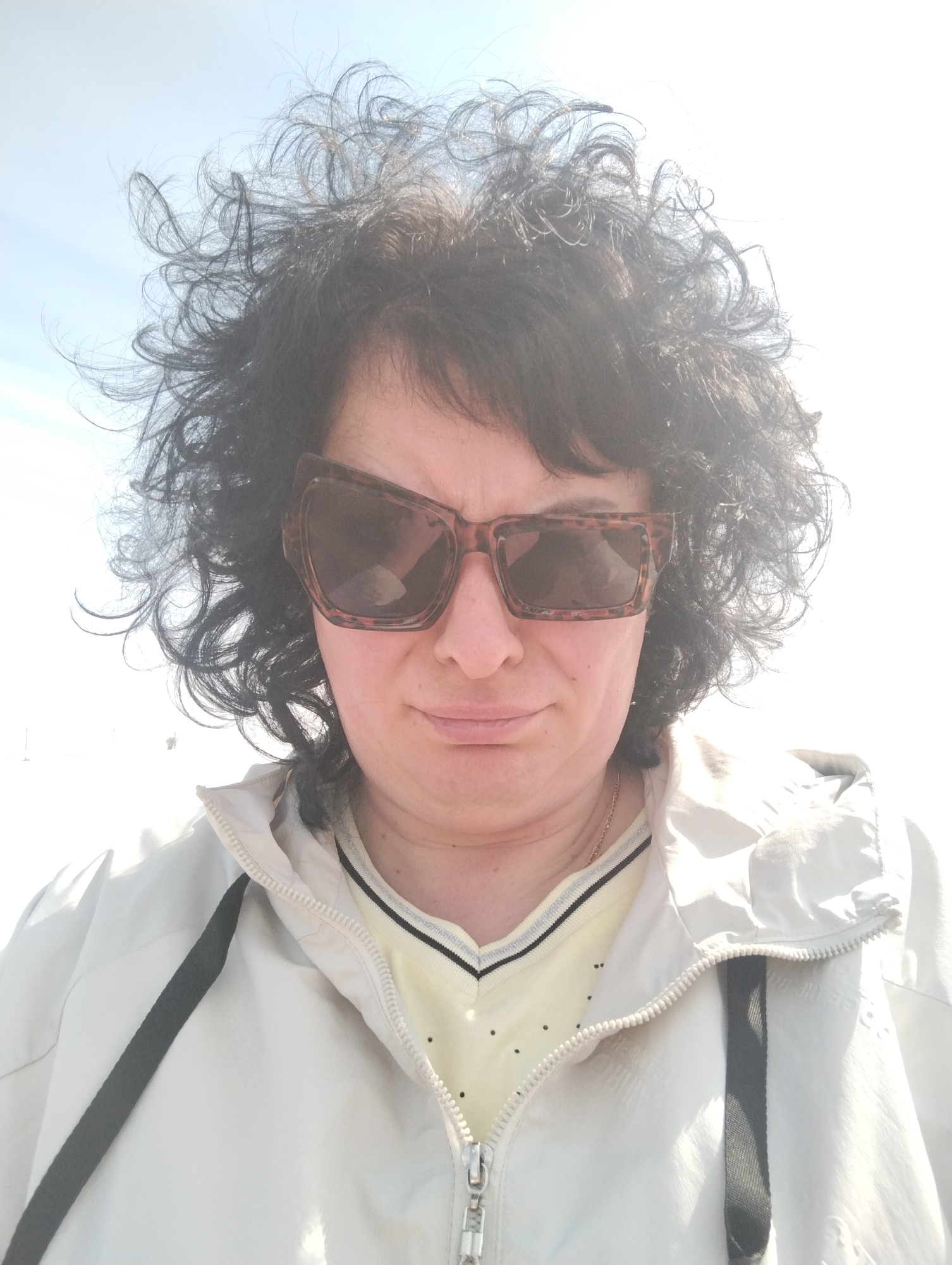 A woman is centered in a high-angle, close-up shot. She has curly, dark hair and wears oversized, patterned sunglasses. Her face is partially obscured by the glasses, but her expression appears neutral, with a slight downturn to her mouth. She is wearing a light-colored jacket with a zipper closure and dark trim, over a yellow top with a V-neckline. The background is washed out by bright sunlight and sky, providing no clear details. No other subjects or objects are visible, and there is no discernible action. The location is unknown.