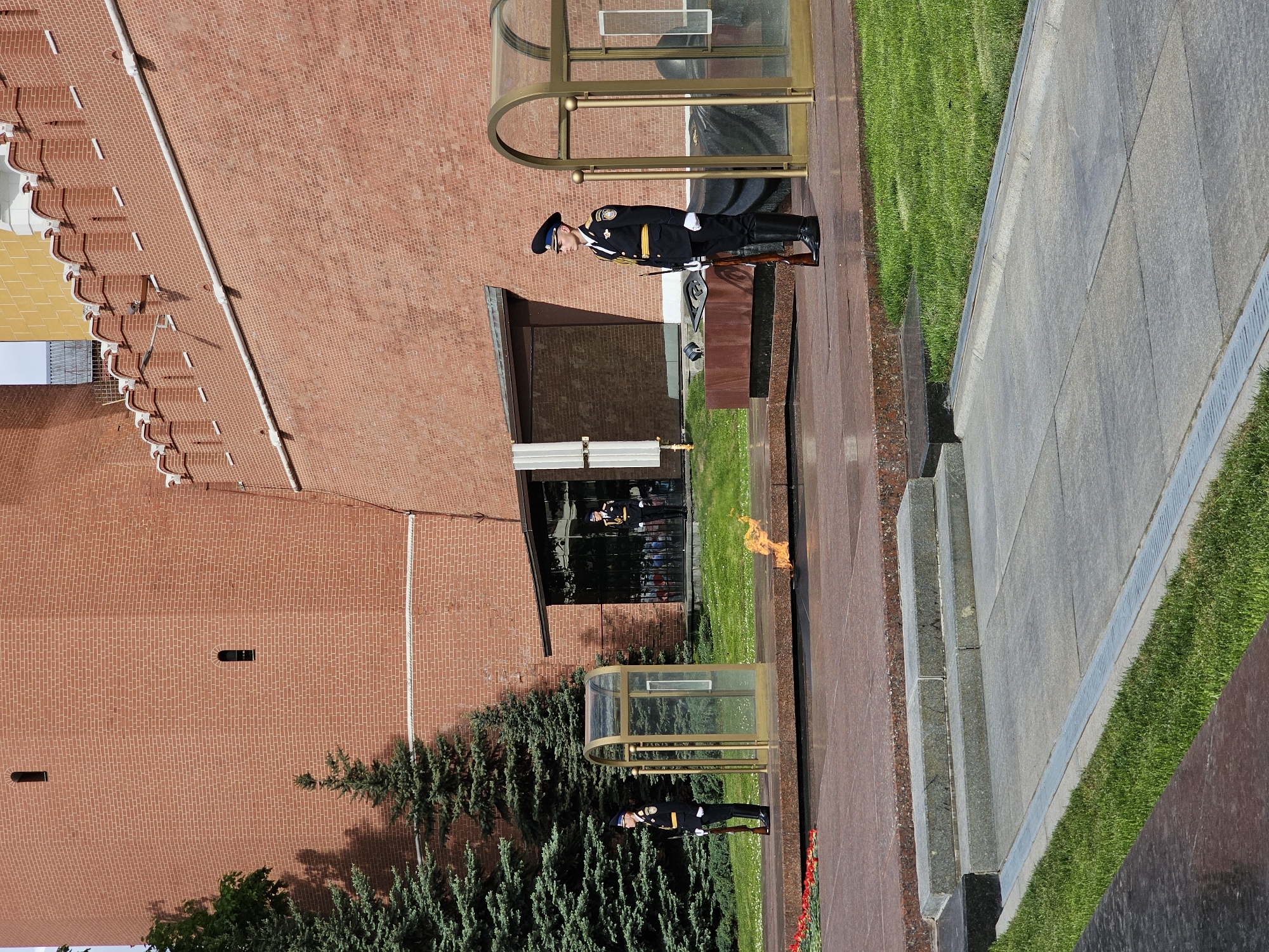 The image shows the Tomb of the Unknown Soldier in Moscow, Russia. A guard in a dark uniform, wearing a ceremonial hat and white gloves, stands at attention with a rifle in hand. Behind the guard is a structure, possibly an enclosure or sentry box, made of glass and a brass-colored metal. To the left of the structure, a flame burns above a memorial. The background includes a red brick wall, likely part of the Kremlin, along with green foliage from trees. A paved area extends into the foreground, with sections of grass and stone steps leading up to the memorial.