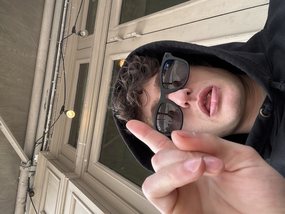 The image captures a male individual in a close-up, selfie-style shot taken outdoors in Vincennes, France. The subject, a young adult with short, dark, curly hair, wears a black hooded sweatshirt with the hood pulled up. He is wearing black rectangular Ray-Ban sunglasses, which reflect indistinct figures and an outdoor scene. His mouth is slightly agape, revealing teeth and a portion of his tongue, on which a small, light-colored particle is visible. His left hand is raised into the foreground, with the index finger extended vertically. The background consists of a light-colored building facade featuring paneled architecture and large windows. Above the facade, a light-grey awning extends. String lights with illuminated, warm-toned bulbs are strung horizontally across the upper part of the frame, attached to the building structure.
