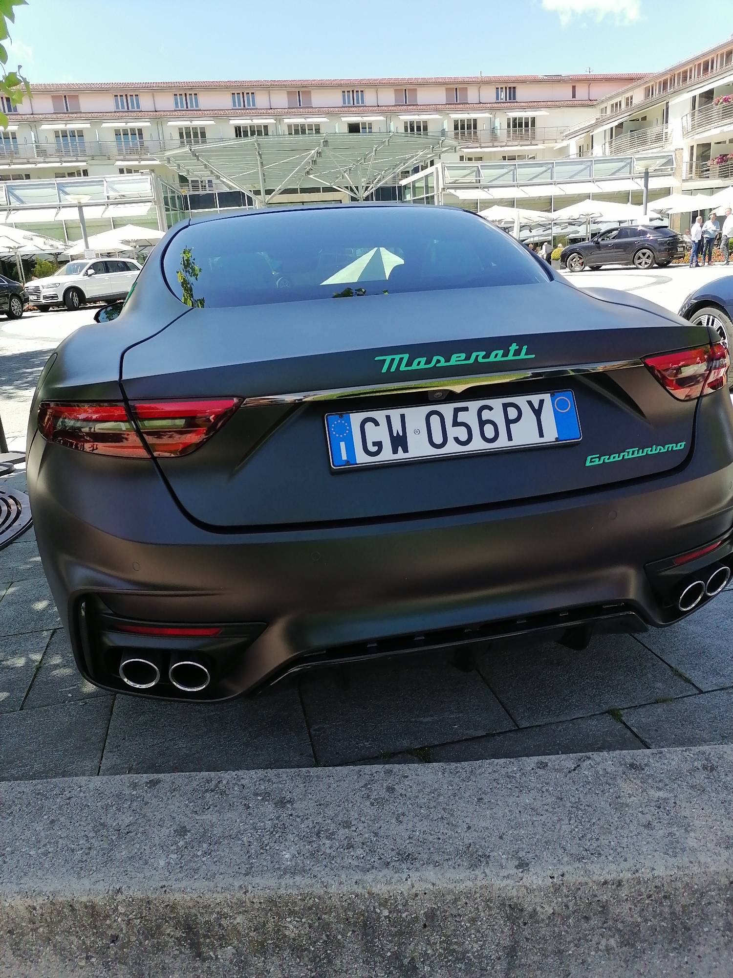 A matte black Maserati GranTurismo with license plate GW 056PY is parked on a paved area in front of a modern building complex. The building has multiple levels with balconies and awnings, and appears to be a hotel or resort. Several other cars, including a white SUV and a dark grey coupe, are visible in the background, as well as a few people standing and walking. The sky is clear and blue with a few wispy clouds. The car's rear is facing the viewer, showing its distinctive taillights, quad exhaust pipes, and the "Maserati" and "GranTurismo" badging in teal. The scene is illuminated by bright sunlight, casting shadows on the ground. This location appears to be the outdoor area of the Bachmair Weissach hotel in Rottach-Egern, Germany.