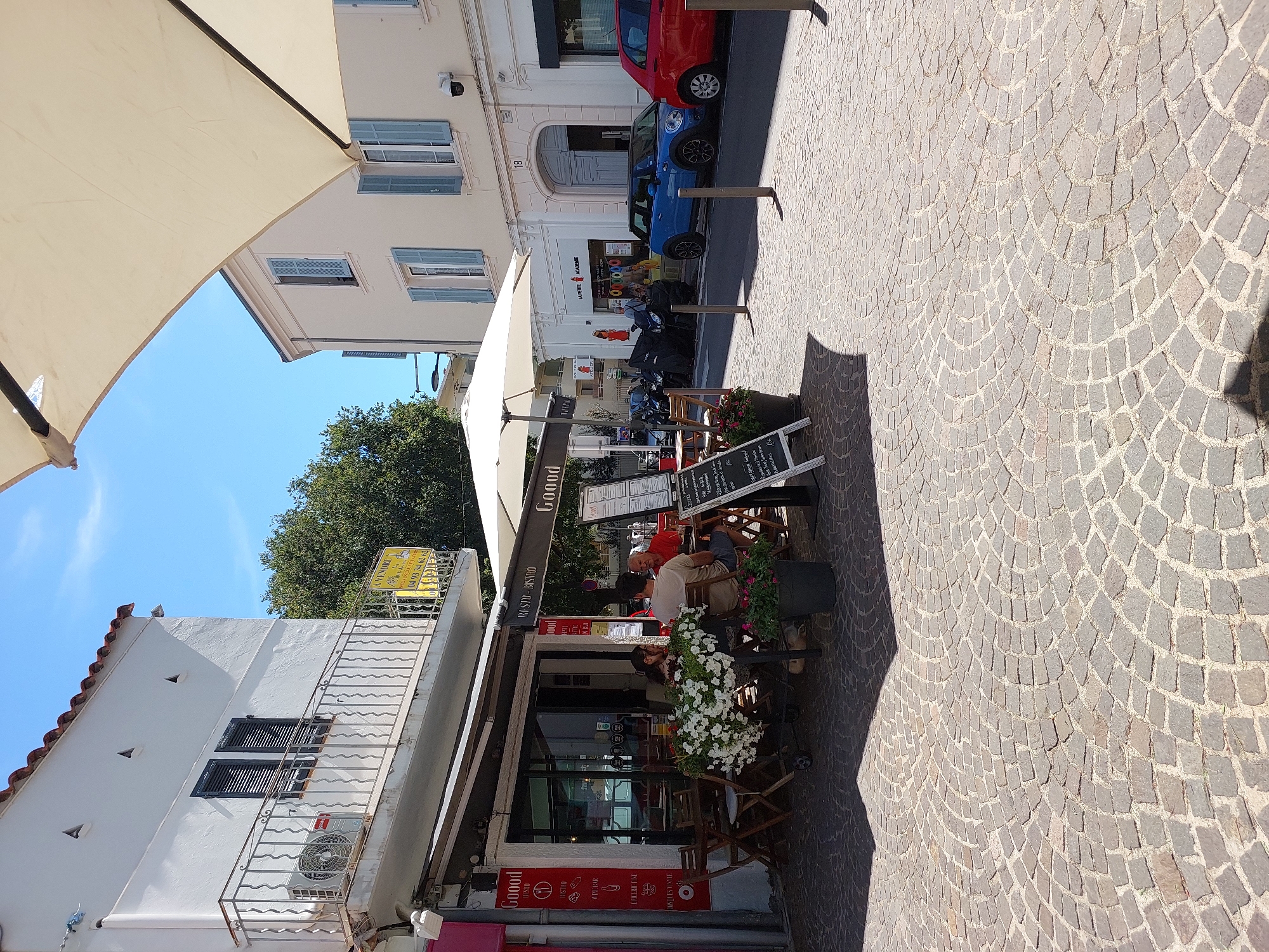 The image depicts a street scene in Antibes, France, likely in the old town area, given the cobblestone pavement and building architecture. In the foreground, a restaurant with outdoor seating is visible. The restaurant's name, "Gooood," is displayed prominently on its signage. Several patrons are seated at tables, enjoying their meal or drinks. A waiter appears to be serving one table. The restaurant has a modern facade with large windows. To the right of the restaurant, a blue Mini Cooper and a red car are parked on the street. The street is paved with traditional cobblestones, and several potted plants and umbrellas add to the ambiance of the outdoor dining area. The buildings in the background are typical of Mediterranean architecture, with white facades and blue shutters. The sky is clear and blue, indicating a sunny day. The scene suggests a pleasant, leisurely atmosphere, with people enjoying the outdoors and the local cuisine.