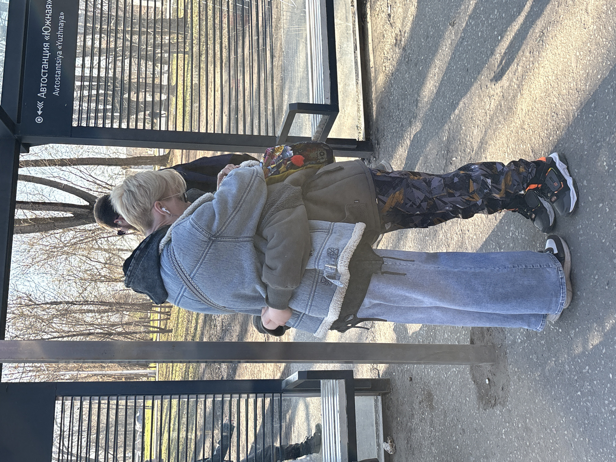 Two individuals are positioned at a bus stop in Perm, Russia. The bus stop signage displays the name "Avtostantsiya «Yuzhnaya»."
One individual is wearing a denim jacket, jeans, and appears to have blonde hair. The second individual is wearing dark camouflage-patterned pants and a jacket with a patterned hat.
The environment is a typical urban setting, including bare trees and a sidewalk. Shadows are cast on the ground, indicating direct sunlight.
