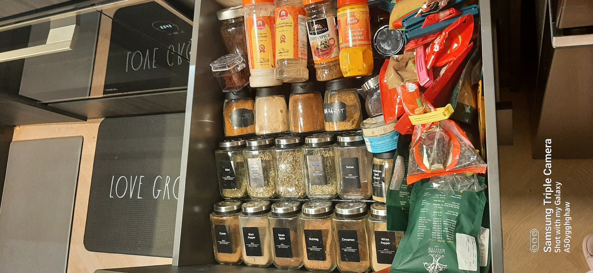 The image shows an open drawer, likely in a kitchen, containing an assortment of spices in glass jars. The drawer also contains a green bag with Arabic text, some colorful plastic clips, and other miscellaneous items. The lighting suggests an indoor environment. The image is watermarked with "Samsung Triple Camera Shot with my Galaxy A50yghghaw", indicating it was taken with a Samsung smartphone. There is no discernible information in the image to suggest a location in Dubai, United Arab Emirates.