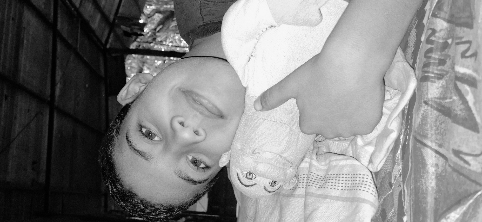A black and white, close-up portrait shot captures a male juvenile, estimated to be pre-adolescent, in an indoor setting within Vallecito, Venezuela. The subject is positioned horizontally, resting his head on a light-colored, soft plush toy with embroidered eyes and mouth. He displays a slight smile and extends his right hand with the thumb raised, indicating a 'thumbs up' gesture. His eyes are directed towards the viewer. The bedding beneath him features a striped and dotted pattern on the pillow and a darker, patterned sheet below. The background is dimly lit, revealing a dark, structured ceiling, possibly made of wood or corrugated material.