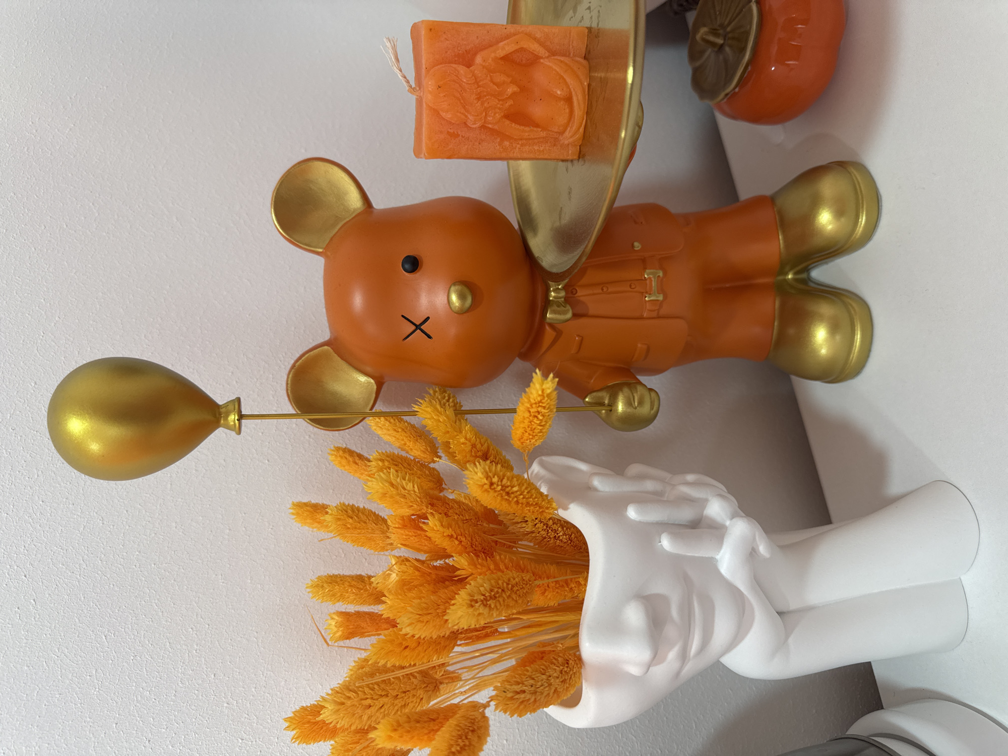 This image presents a vibrant and carefully arranged still life composition of decorative objects, set indoors against a plain white, textured wall. The dominant color scheme features warm oranges and golds, accented by white.
Central to the display is an orange figurine resembling a teddy bear, featuring golden ears, a golden nose, and golden feet. Its face is simplified with a small black dot for one eye and a black 'X' for a mouth. The figurine wears a golden backpack or satchel on its back. In its left hand, it holds a golden balloon on a thin rod, while its right hand supports a small, reflective golden tray.
Upon this golden tray rests an orange, rectangular candle. It is embossed with a detailed relief of a reclining or dynamic female figure, possibly a mermaid or classical goddess, identifiable by her flowing hair.
In the upper right corner of the background, part of another orange decorative object with golden accents is visible, resembling a small fruit such as a persimmon.
At the bottom right, a distinctive white vase is shaped like two hands cupped upwards. This vase is filled with a dense arrangement of dried, bright yellow-orange grass-like flowers, characterized by their fluffy, elongated seed heads, likely a type of ornamental dried grass such as lagurus (bunny tail grass).
The overall impression is one of a decorative, whimsical, and perhaps autumnal or luxurious display. No activity is taking place, and there is no visible text. The location context provided for this image is Sochi, Russia.