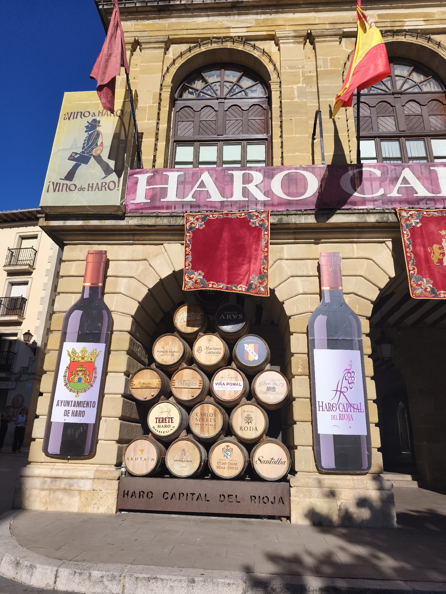 The image depicts a festive outdoor scene in the city of Haro, Spain, likely during a wine-related event. In the foreground, a large, stylized advertisement of a wine bottle stands to the left, featuring a label with the "Ayuntamiento de Haro" (City Council of Haro) emblem and text. To its right, a pyramid of wine barrels is displayed, each bearing different winery labels and names such as "ARVS," "BODEGAS," "RODA," "MUGAS," "AKUTAIN," "MANZANOS," and "SANTALBA." Above the barrels, two large banners are hung, one with the text "HARO CAPITAL" and another featuring the Spanish flag and red flags. The background consists of a building with arched windows and stone architecture. Shadows suggest it is a sunny day. Two women are visible in the background on the left side of the image, near a building with balconies.