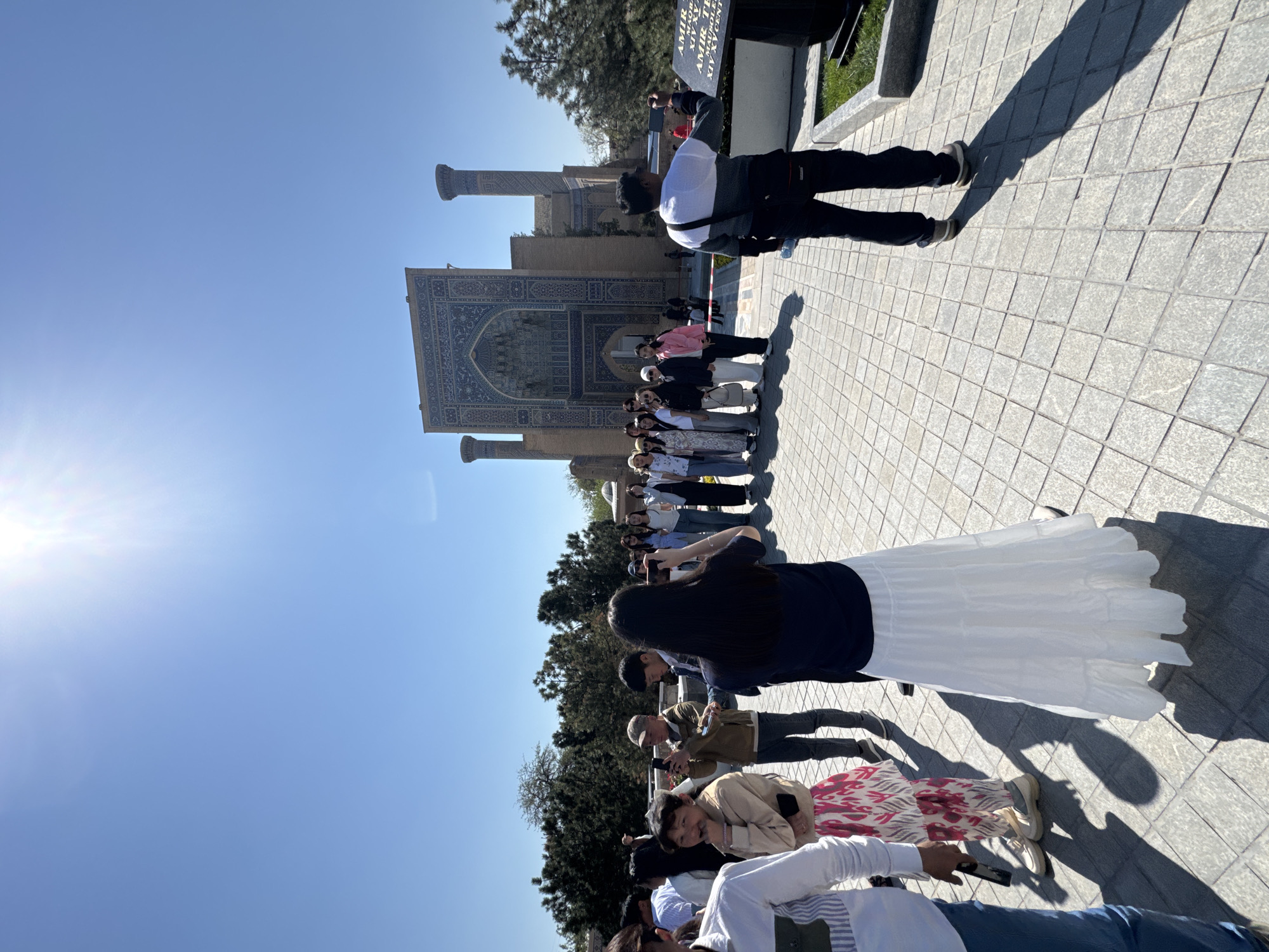 A bright outdoor scene shows tourists in front of the Gur-e-Amir Mausoleum in Samarkand City, Uzbekistan. The building has a large, decorated archway flanked by two minarets. The foreground is filled with people, many holding phones and taking pictures. A man in a white shirt and dark pants stands close to a tombstone-like structure, possibly a memorial. The ground is paved with light-colored square tiles. The sky is clear and bright blue.