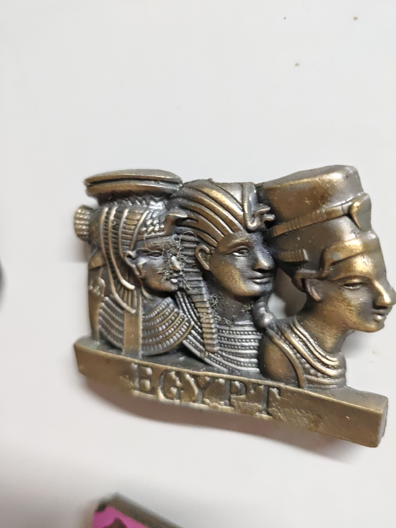 A close-up shot depicts a bronze-colored souvenir magnet with three Egyptian figures in profile. The word "EGYPT" is inscribed on the base of the magnet. The figures are adorned with traditional Egyptian headdresses and collars. The background is a plain, light-colored surface.
The location of the image cannot be determined with the available information.