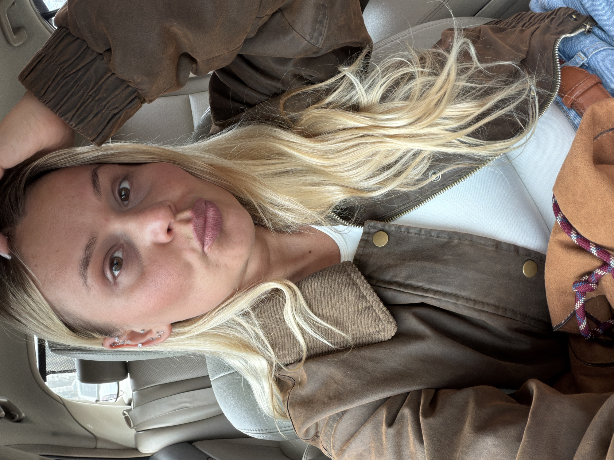 The image shows a selfie of a blonde woman, likely inside a car. The woman has fair skin, light blonde hair, and several piercings in her ear. She is wearing a brown jacket over a white top. The car's interior is visible, including the grey leather seat. The image does not provide sufficient detail to determine if the location is the city of Sarapul, Russia.