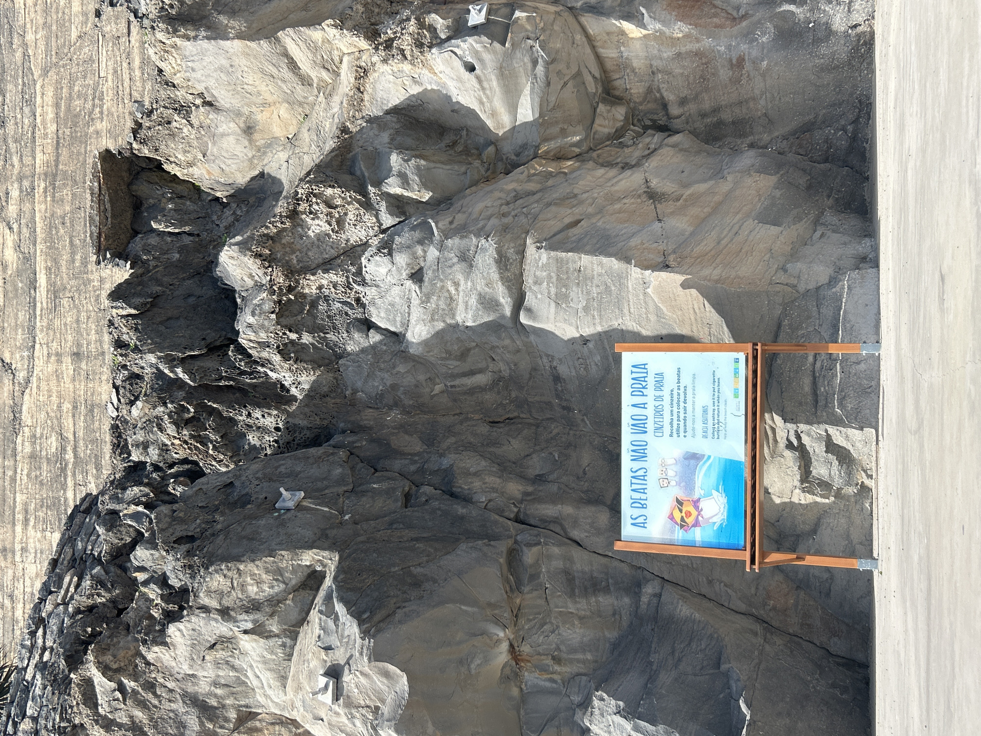 The image shows a sign that reads "AS BEATAS NÃO VÃO À PRAIA" (Cigarette butts don't go to the beach) located in Câmara de Lobos, Portugal. The sign is mounted on a wooden frame and is placed against a rocky cliff. The sign has instructions on how to collect and dispose of cigarette butts in beach ashtrays. The environment is rocky and likely near a beach or coastal area. There is also a concrete wall or structure on one side of the image.
