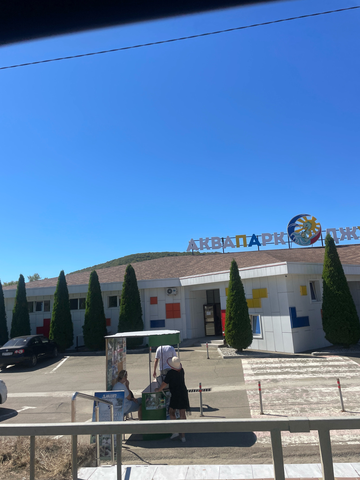 The image shows the entrance to a water park in Dzhubga, Russia. The water park's name, "АКВАПАРК ДЖУБГА," is visible in large, colorful letters on the building. The building itself is white with colorful rectangular accents. Several tall, green trees line the front of the building. In the foreground, there is a parking lot with a black car. Near the entrance, a kiosk with information displayed on it is set up, and several people are gathered around it. One person is seated, while another appears to be interacting with someone behind the kiosk. A third person, wearing a white shirt and a hat, is also near the kiosk. A pedestrian crossing is visible in front of the building. The sky is a clear blue.