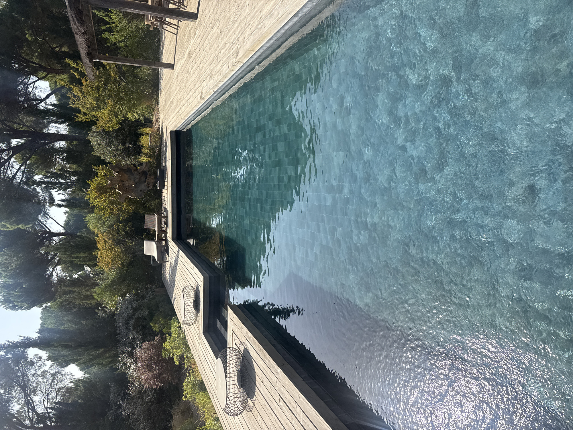 This image depicts a private swimming pool area in Grândola, Portugal. The scene is outdoors, with a focus on a rectangular swimming pool with turquoise-tiled lining, surrounded by a wooden deck. The pool is illuminated by natural sunlight, casting reflections on its surface. Two black wire mesh tables with intricate designs are placed on the wooden deck. In the background, lush greenery and trees are visible, creating a natural and serene environment. To the right of the pool, a wooden pergola with a thatched roof shelters a dining table and chairs. There are no people visible in the image. The overall atmosphere is tranquil and inviting, suggesting a leisure space.