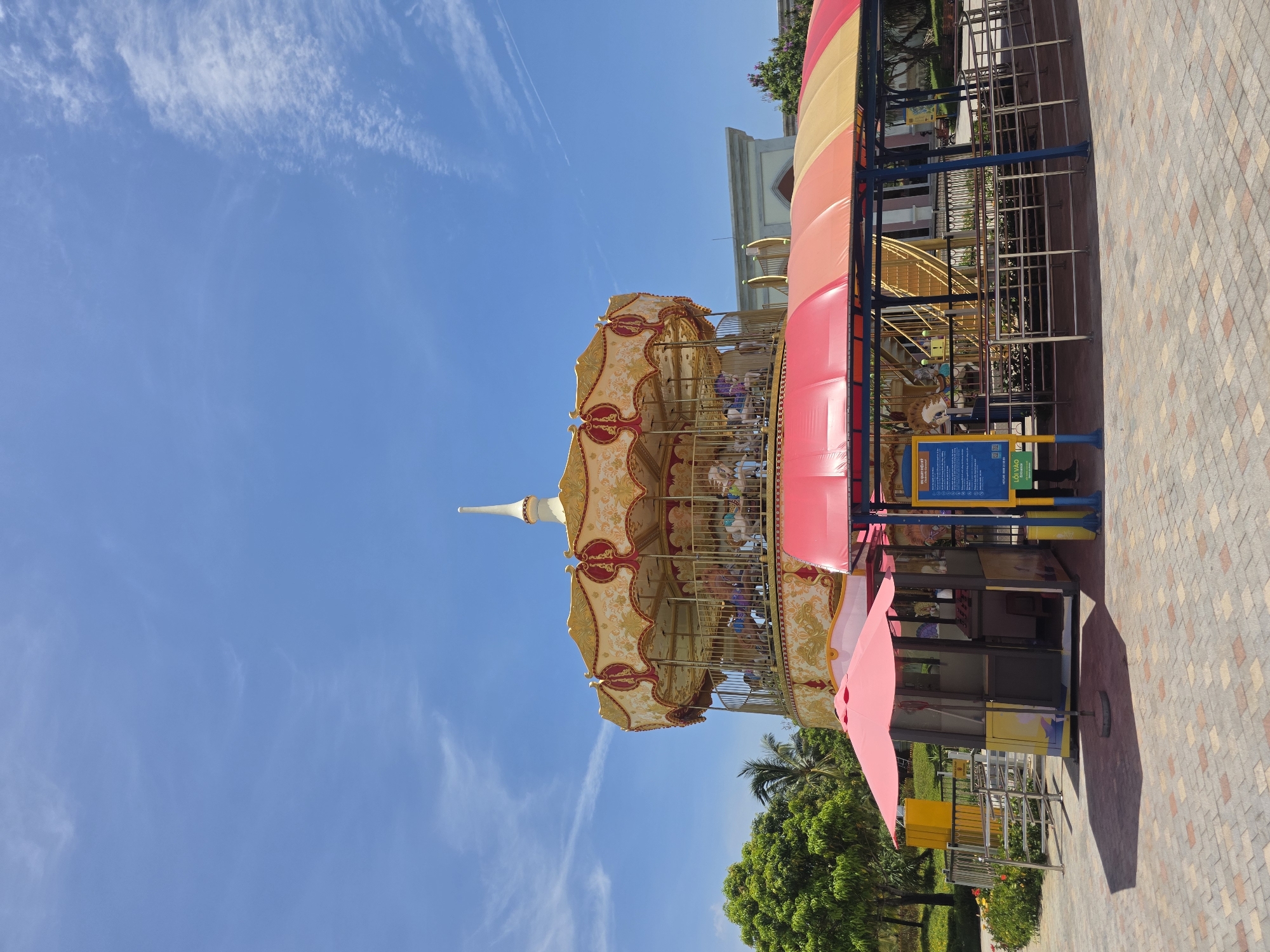 The image depicts a carousel located outdoors in Đà Nẵng, Vietnam. The carousel is predominantly gold and red, with a conical roof and a spire at the top. People are visible riding the carousel. A striped canopy is adjacent to the carousel, providing shade. A metal fence surrounds the carousel entrance. A sign with text in Vietnamese is attached to the fence. The ground is paved with bricks. In the background, trees and a building are visible. The sky is blue with scattered clouds.