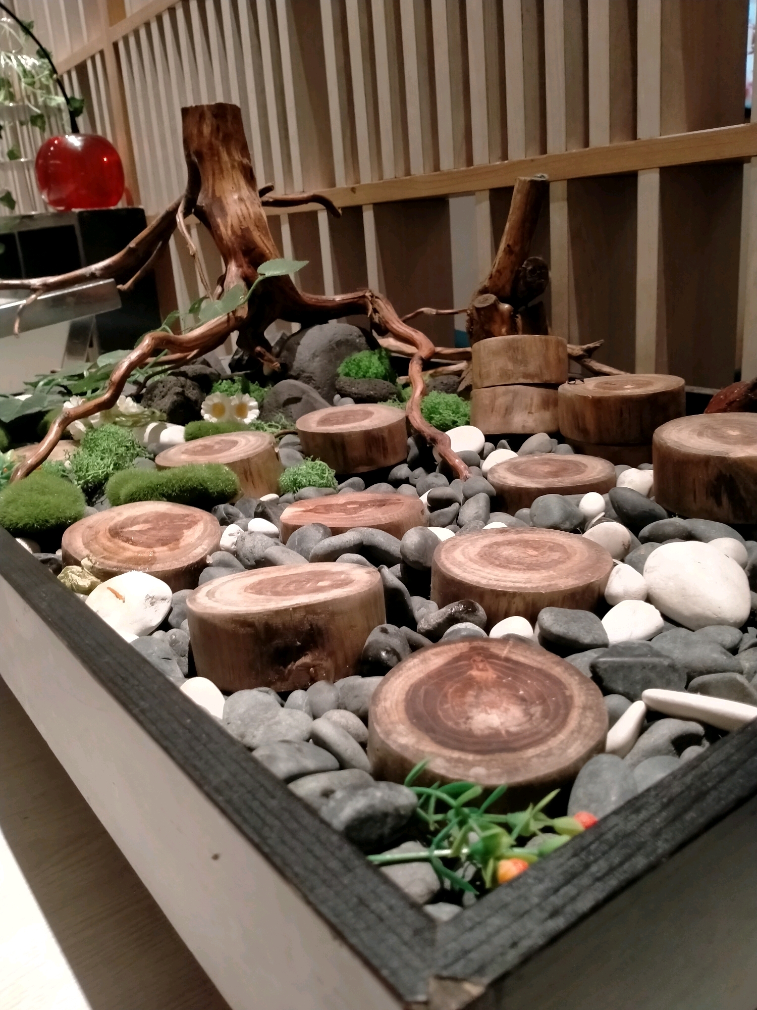 This image captures a decorative arrangement within what appears to be a restaurant or cafe setting, possibly located in Batu, Indonesia, suggested by the presence of natural elements commonly found in that region. The foreground features a planter box filled with dark gray and white pebbles. Several circular wooden slices, showcasing the tree rings, are strategically placed on top of the pebbles. Some of these wooden slices are stacked. Interspersed among the pebbles and wooden elements are small, vibrant green mossy mounds and two delicate white artificial flowers. In the background, a substantial piece of driftwood with intricate branches is prominently displayed, standing upright. Behind the driftwood, a vertical slatted wooden panel creates a textured backdrop. To the left of the driftwood, a bright red, spherical object, possibly a decorative glass ornament, rests on a dark surface. The overall scene evokes a natural, zen-like aesthetic, commonly seen in establishments aiming for a tranquil ambiance. The lighting appears to be warm and ambient, highlighting the textures and colors of the natural materials.