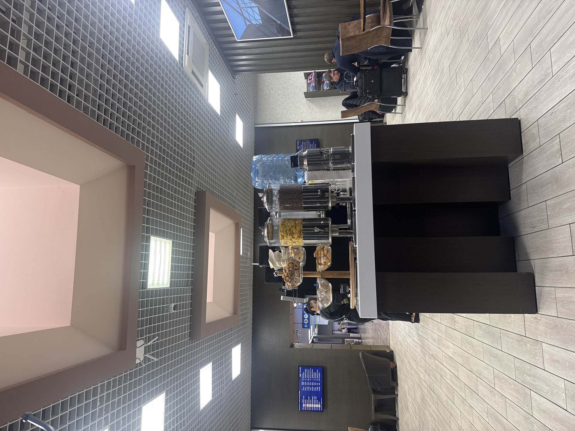 The image shows an airport lounge. Visible are lounge chairs, a water/coffee dispenser with snacks, a schedule board, and people. The location is likely Sochi, Russia, given the tags.
