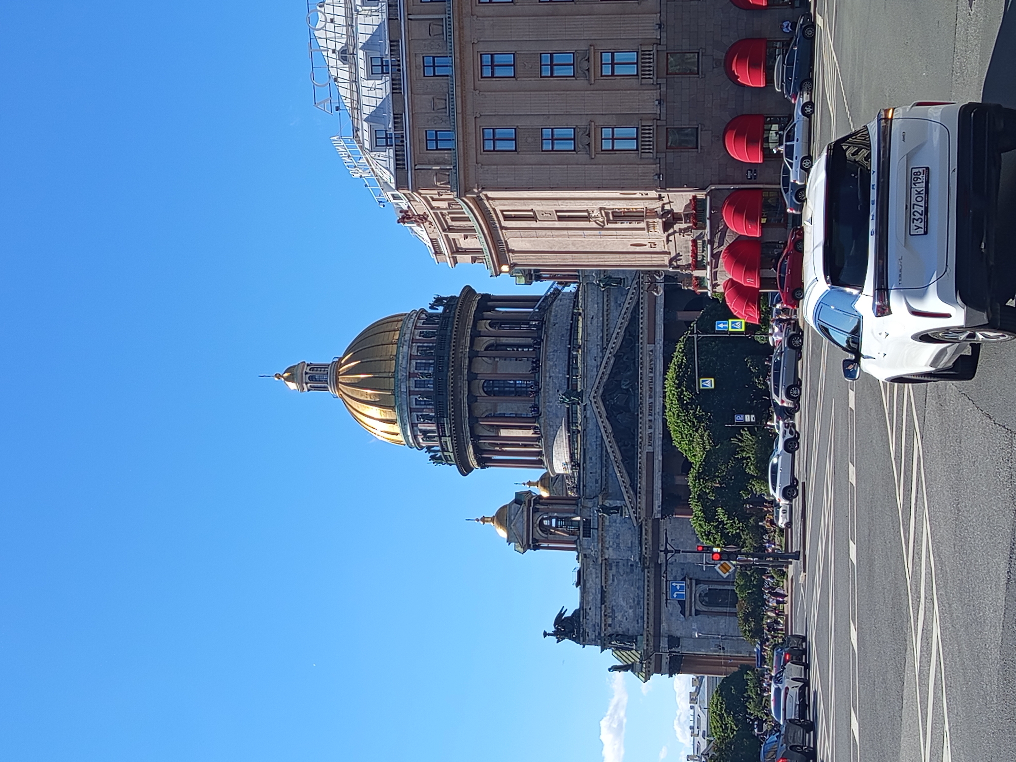 The image captures a street-level view of St. Isaac's Cathedral in Saint Petersburg, Russia, on a clear, sunny day. The grand cathedral, with its prominent golden dome and neoclassical architecture, dominates the background. To the right of the cathedral, a light-colored, multi-story building with arched windows and red awnings is visible. Several cars are present on the street, including a white SUV in the foreground. Pedestrians are walking on the sidewalks and crossing the street in the middle ground. The scene is set at an intersection with traffic lights and road markings. The sky is a vibrant blue, with a few wispy clouds.