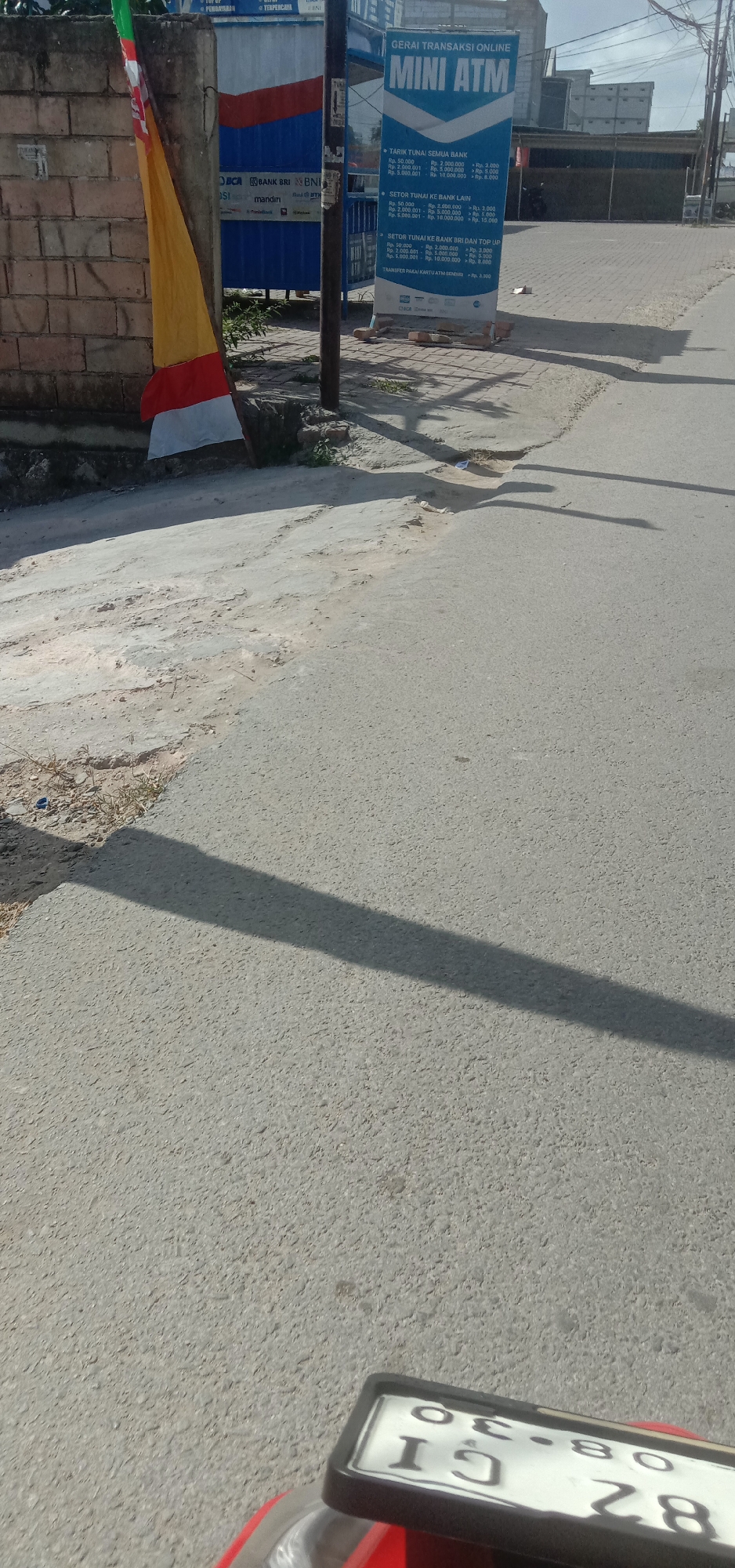 The image captures a street scene in Kendari, Indonesia, likely near a financial institution or business district. In the foreground, a portion of a red motorcycle with a license plate bearing the characters "OG 30 IG 28" is visible. The road surface is asphalt, showing some wear and tear with patches of lighter-colored material. A long shadow of what appears to be a lamppost or utility pole stretches across the road.
To the left, a red brick wall stands, partially obscured by a flag with red and white stripes. Adjacent to the wall is a blue structure with various bank logos, including BRI, Mandiri, and BNI. A blue and white banner advertising "GERAI TRANSAKSI ONLINE MINI ATM" is prominently displayed, listing transaction fees for withdrawing, depositing, and topping up various banks. The banner is propped up by what look like bricks. In the background, buildings and electrical poles are visible under a clear sky. The environment suggests an urban or commercial area with typical street infrastructure.