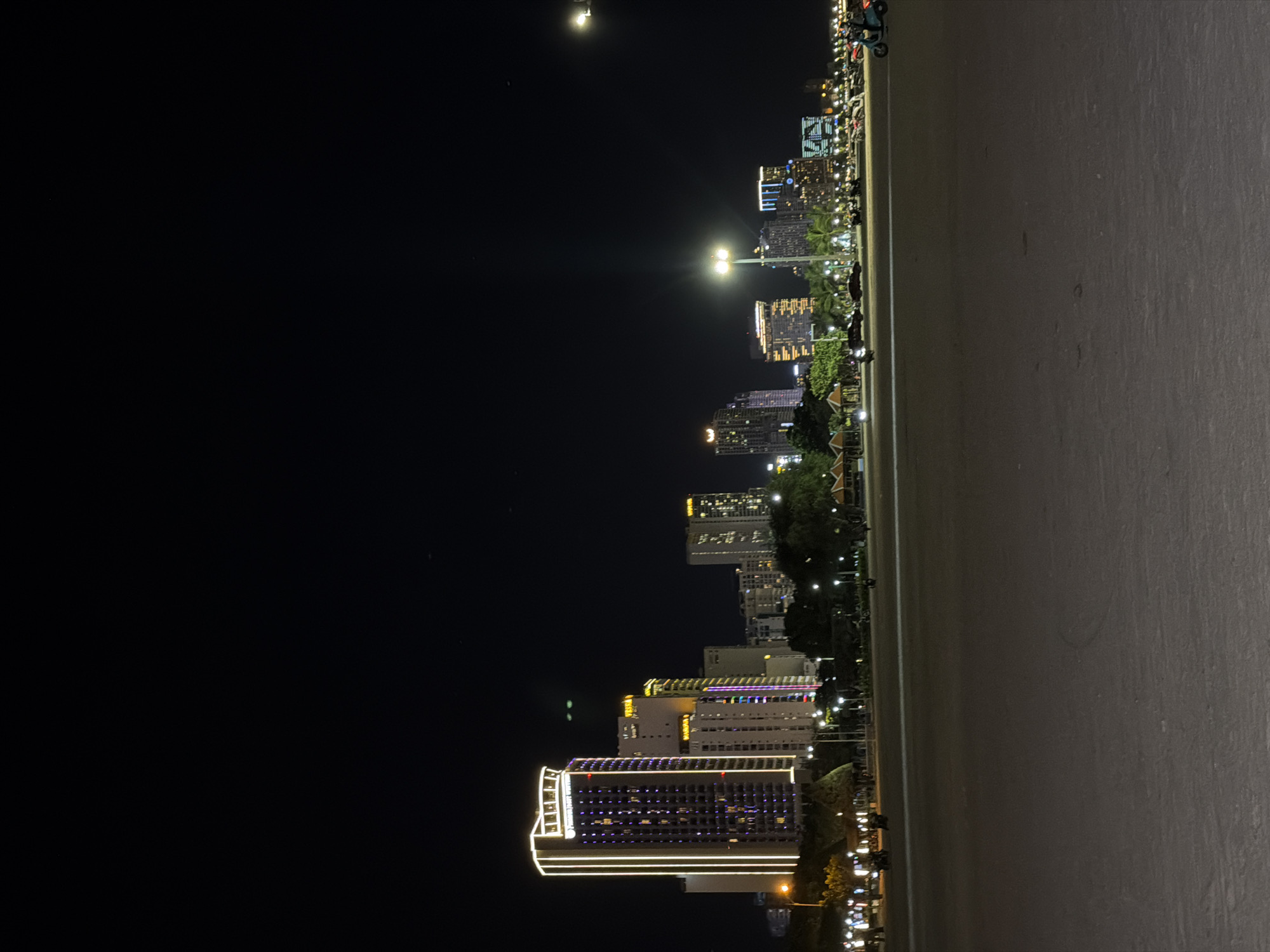 The image shows a night scene in Nha Trang Ward, Vietnam, featuring a cityscape with several illuminated buildings against a dark sky. A bright moon is visible in the sky. The buildings are of varying heights and designs, with some displaying distinct architectural lighting. A paved area, possibly a walkway or sidewalk, runs along the right side of the frame. There are trees and other vegetation interspersed among the buildings, and streetlights provide additional illumination. The scene appears to be from a high vantage point, offering a wide view of the city at night.