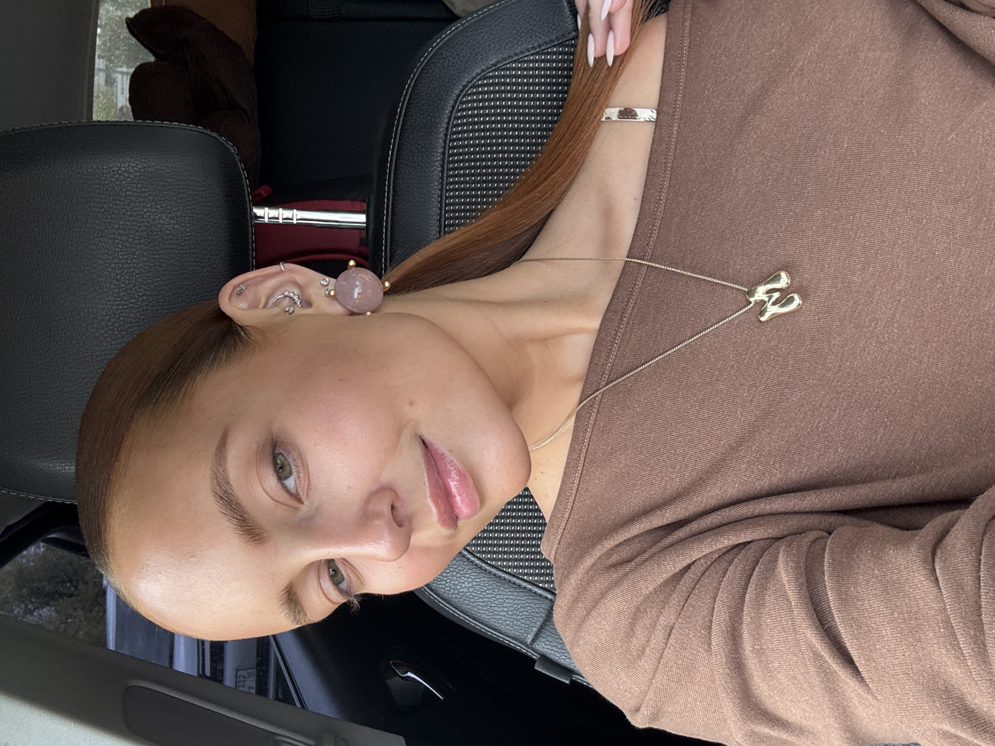 A woman is pictured indoors, likely in a car based on the presence of car seats and interior trim. She is wearing a brown top, gold necklace with an "H" pendant, and several earrings in her left ear, including a pink sphere. Her hair is reddish-brown and pulled back. The car interior appears to be black leather. The image provides no evidence to suggest the location of Chaikovsky, Russia.